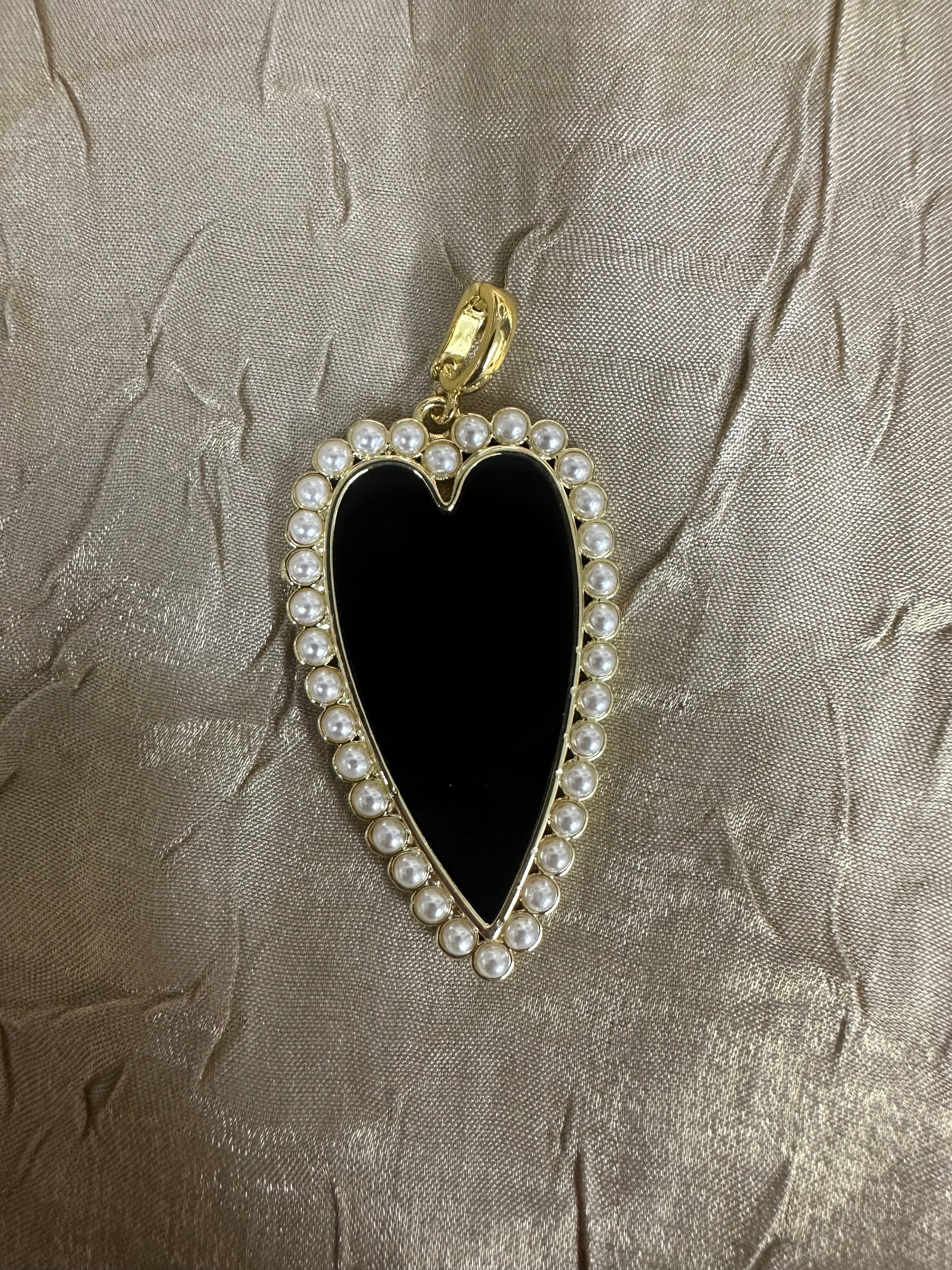 A heart-shaped pendant featuring a glossy black center, bordered by small white faux pearls and a gold-tone metal frame, resting on a textured beige fabric background.