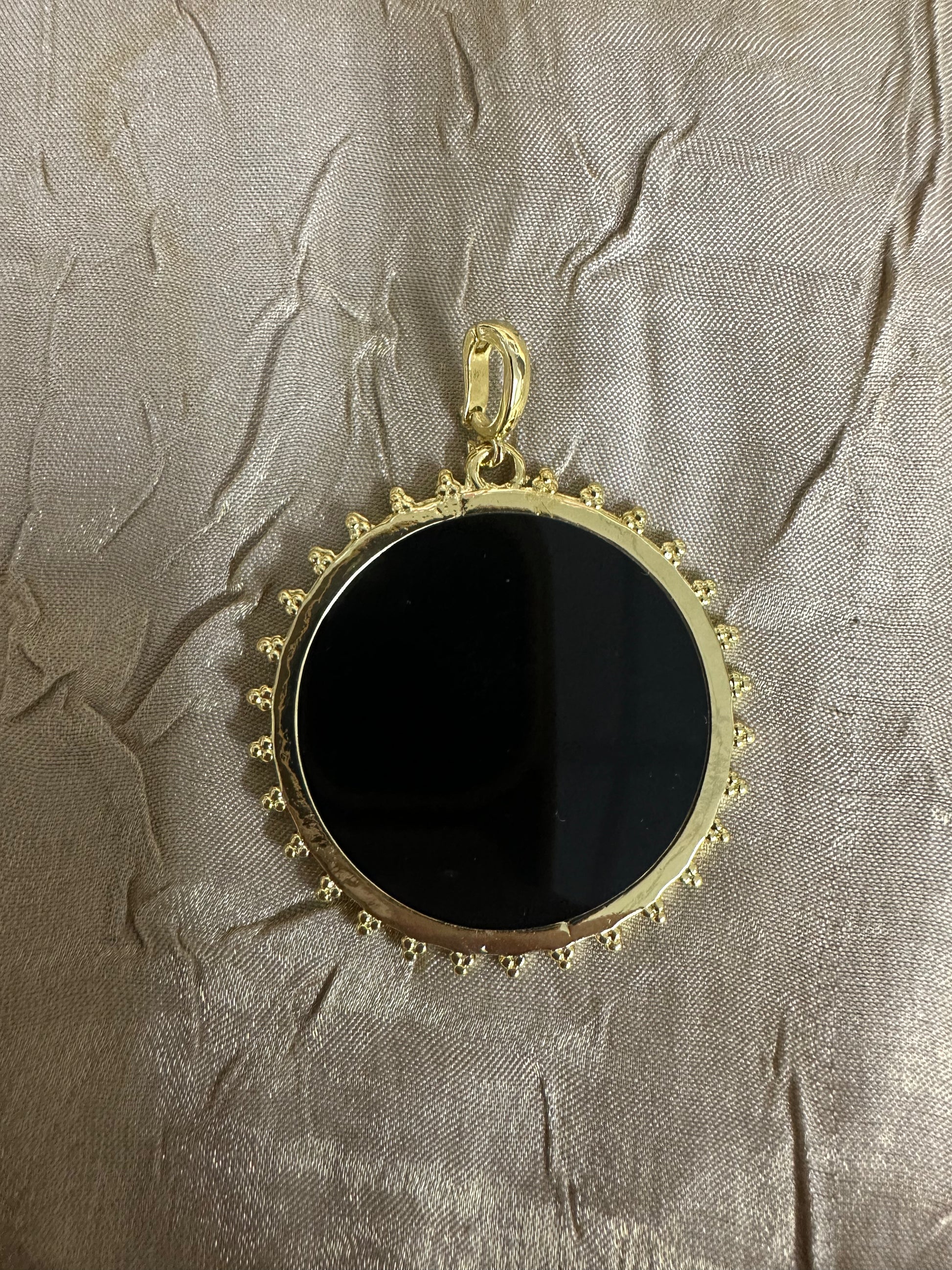 A round black pendant with a gold-tone beaded border and top loop, displayed on a textured beige fabric background.