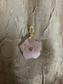 A pink rose quartz flower pendant charm with a gold-plated clasp and a small central clear stone, displayed on a textured beige fabric background.