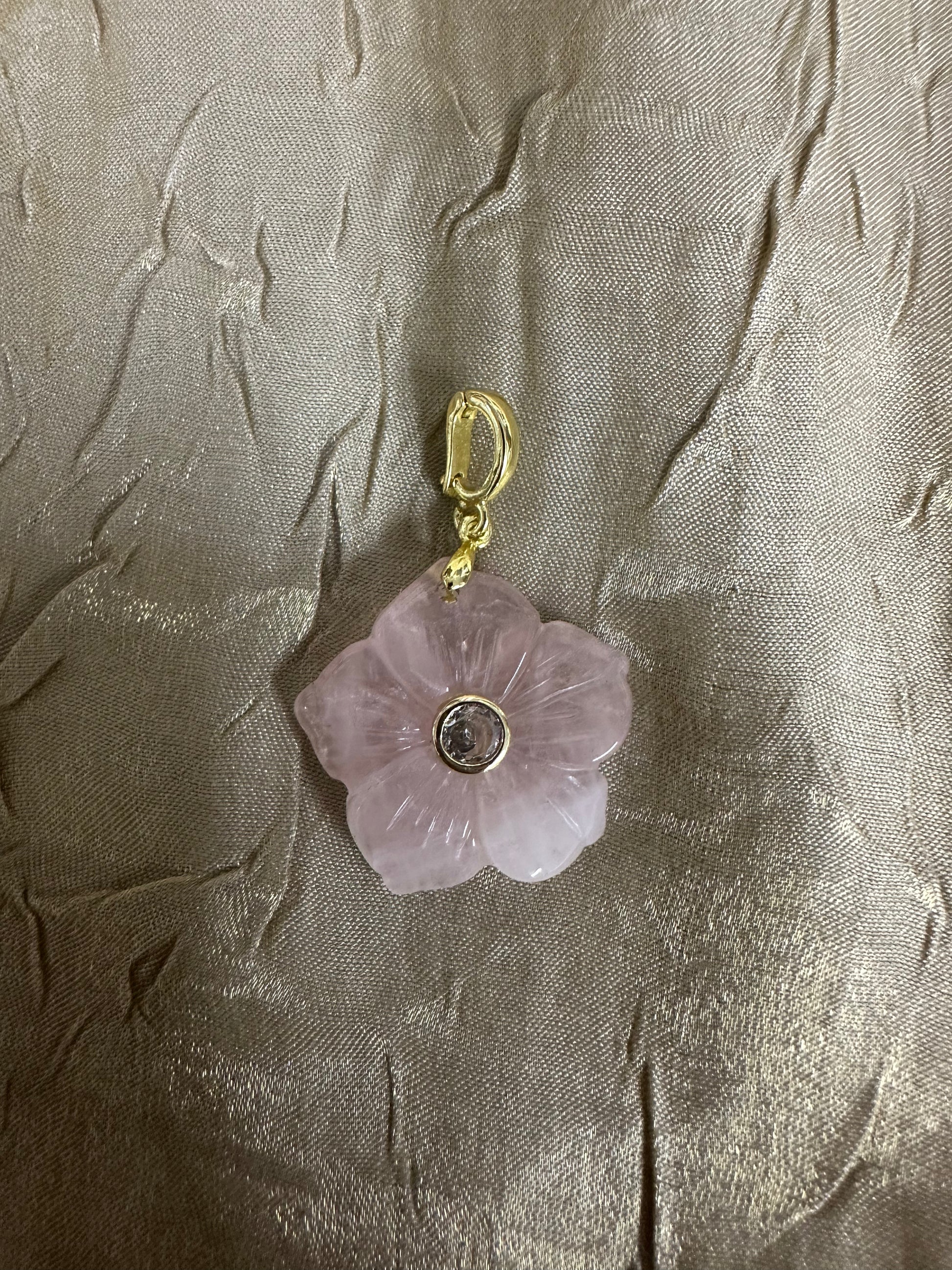 A pink rose quartz flower pendant charm with a gold-plated clasp and a small central clear stone, displayed on a textured beige fabric background.