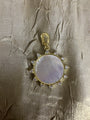 A gold pendant featuring a round mother-of-pearl centerpiece with a halo of small clear stones, displayed on a textured beige fabric background.