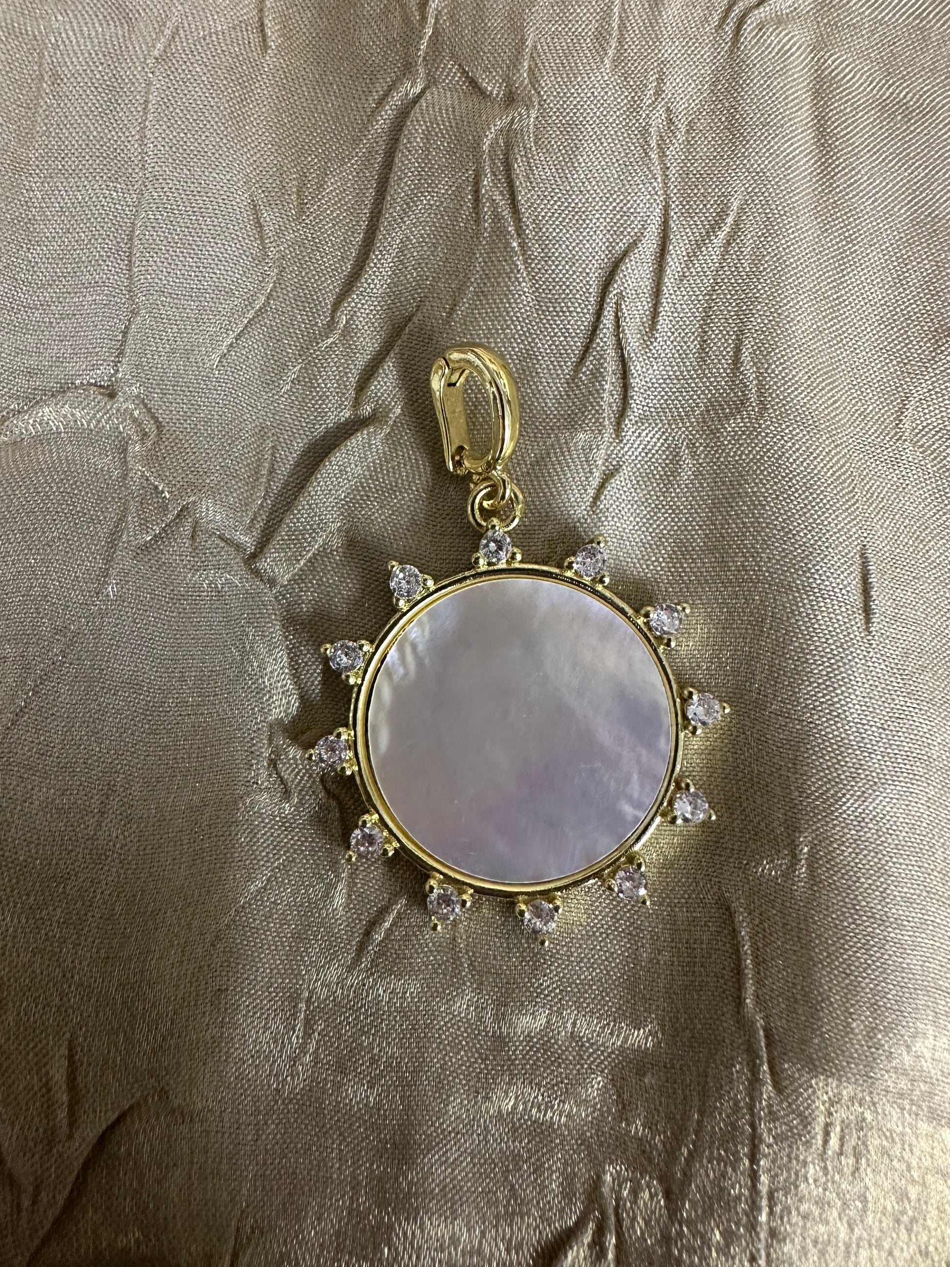 A gold pendant featuring a round mother-of-pearl centerpiece with a halo of small clear stones, displayed on a textured beige fabric background.
