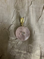 A round pale pink stone pendant with a small clear center stone and a gold-tone bail, photographed on a textured metallic taupe fabric background.