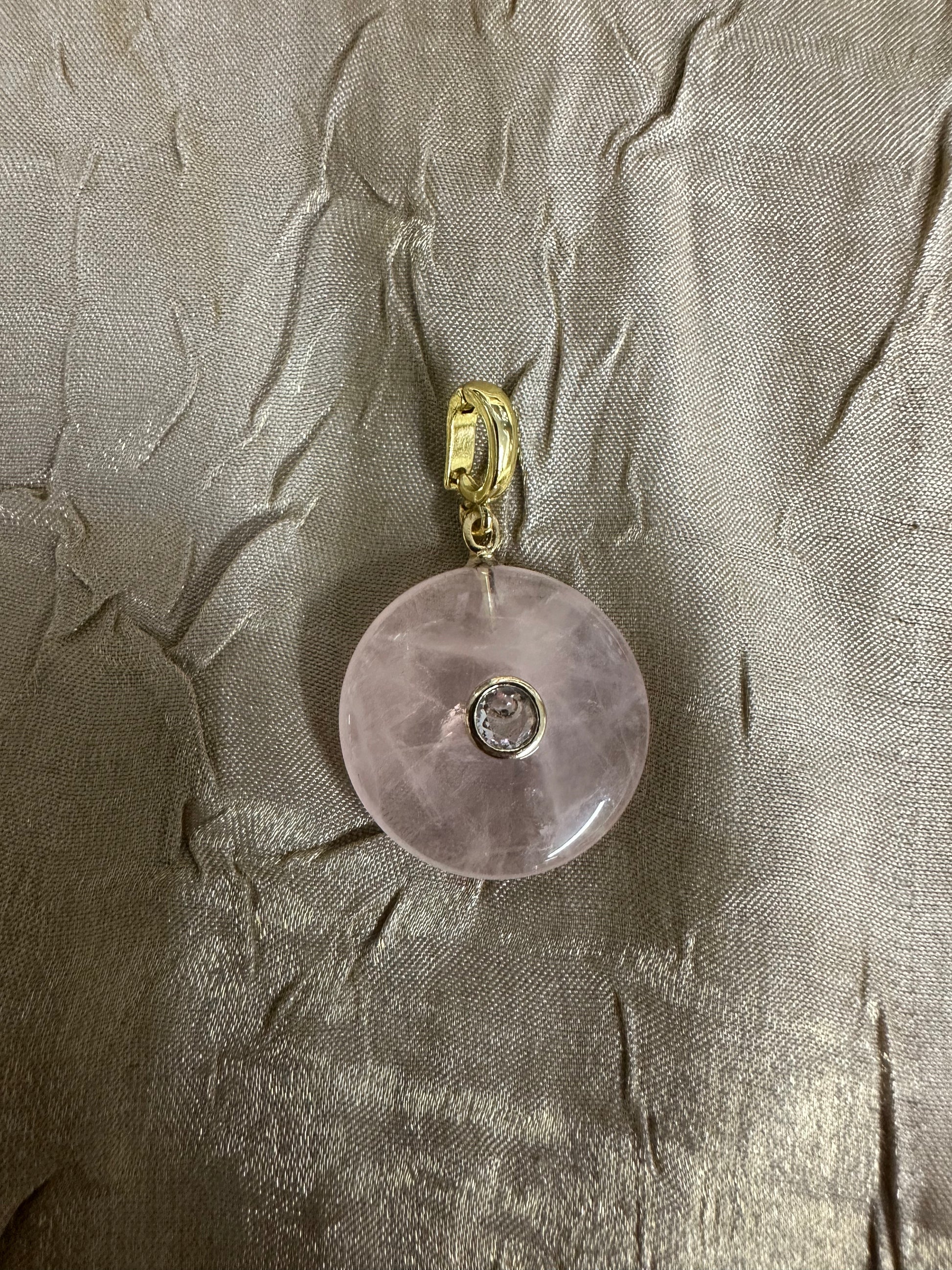 A round pale pink stone pendant with a small clear center stone and a gold-tone bail, photographed on a textured metallic taupe fabric background.