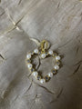 A gold heart-shaped charm or pendant featuring white faux pearls and clear rhinestones, displayed on a textured beige fabric background.