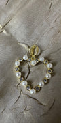 A gold heart-shaped charm or pendant featuring white faux pearls and clear rhinestones, displayed on a textured beige fabric background.