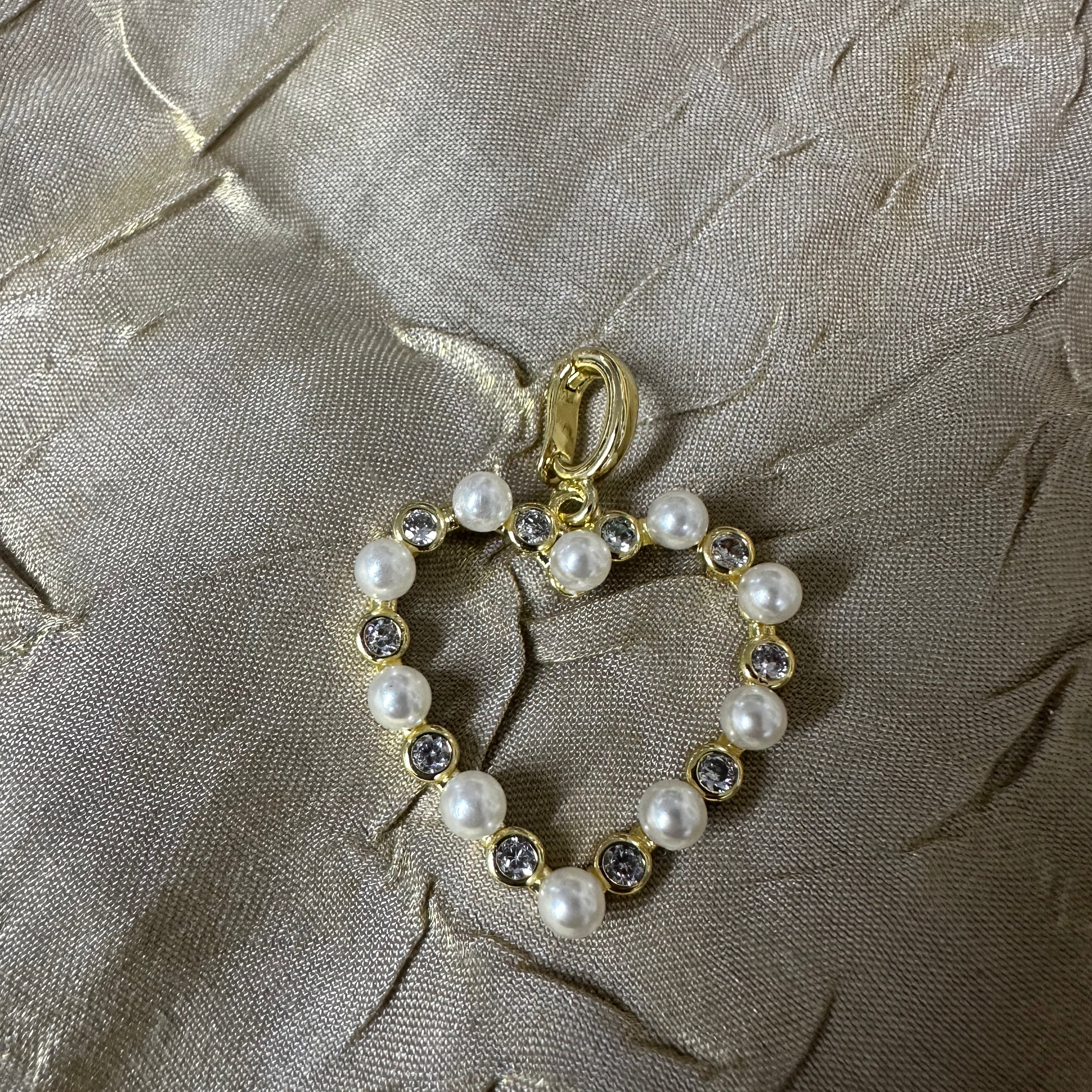 A gold heart-shaped charm or pendant featuring white faux pearls and clear rhinestones, displayed on a textured beige fabric background.