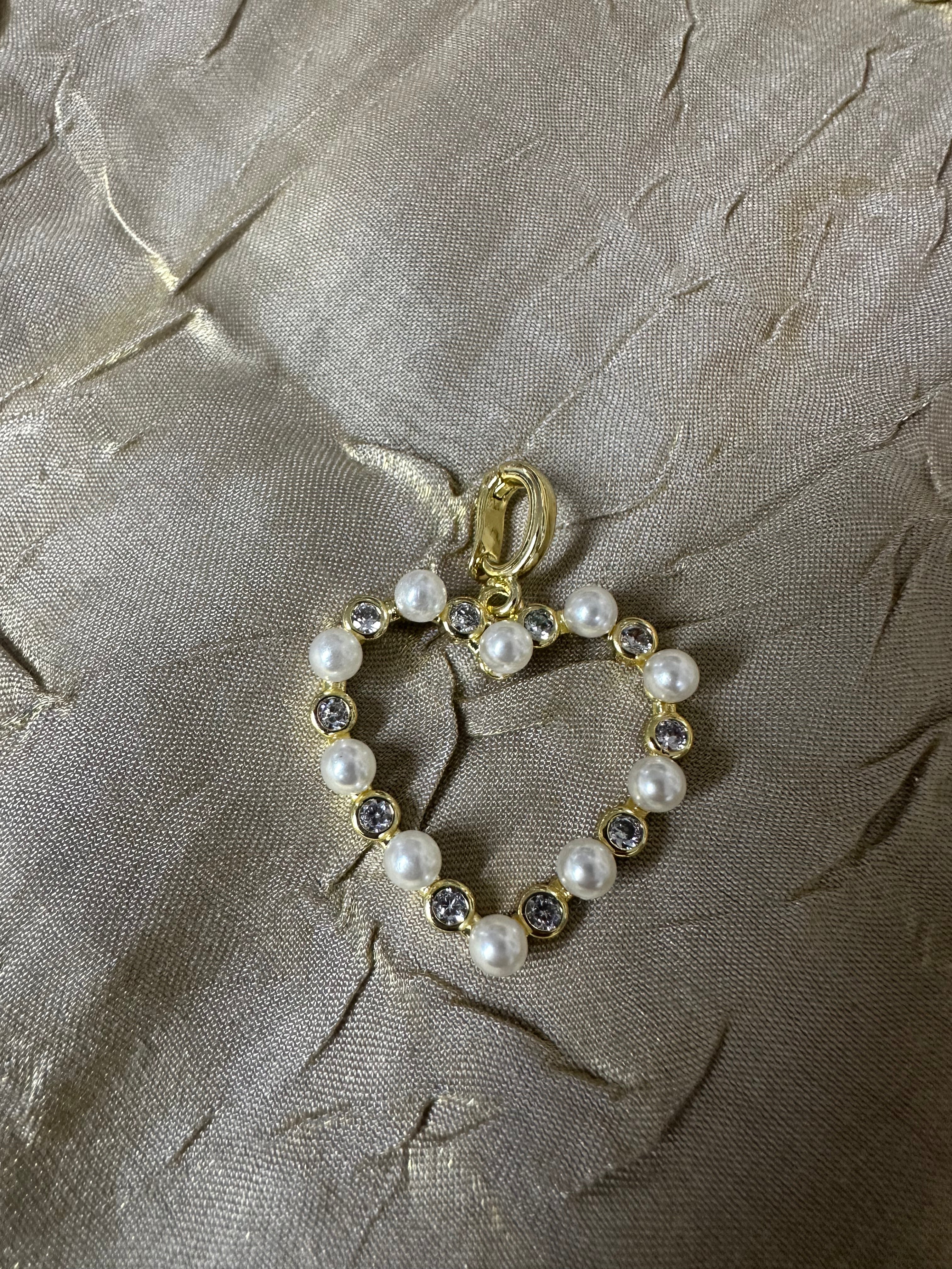 A gold heart-shaped charm or pendant featuring white faux pearls and clear rhinestones, displayed on a textured beige fabric background.