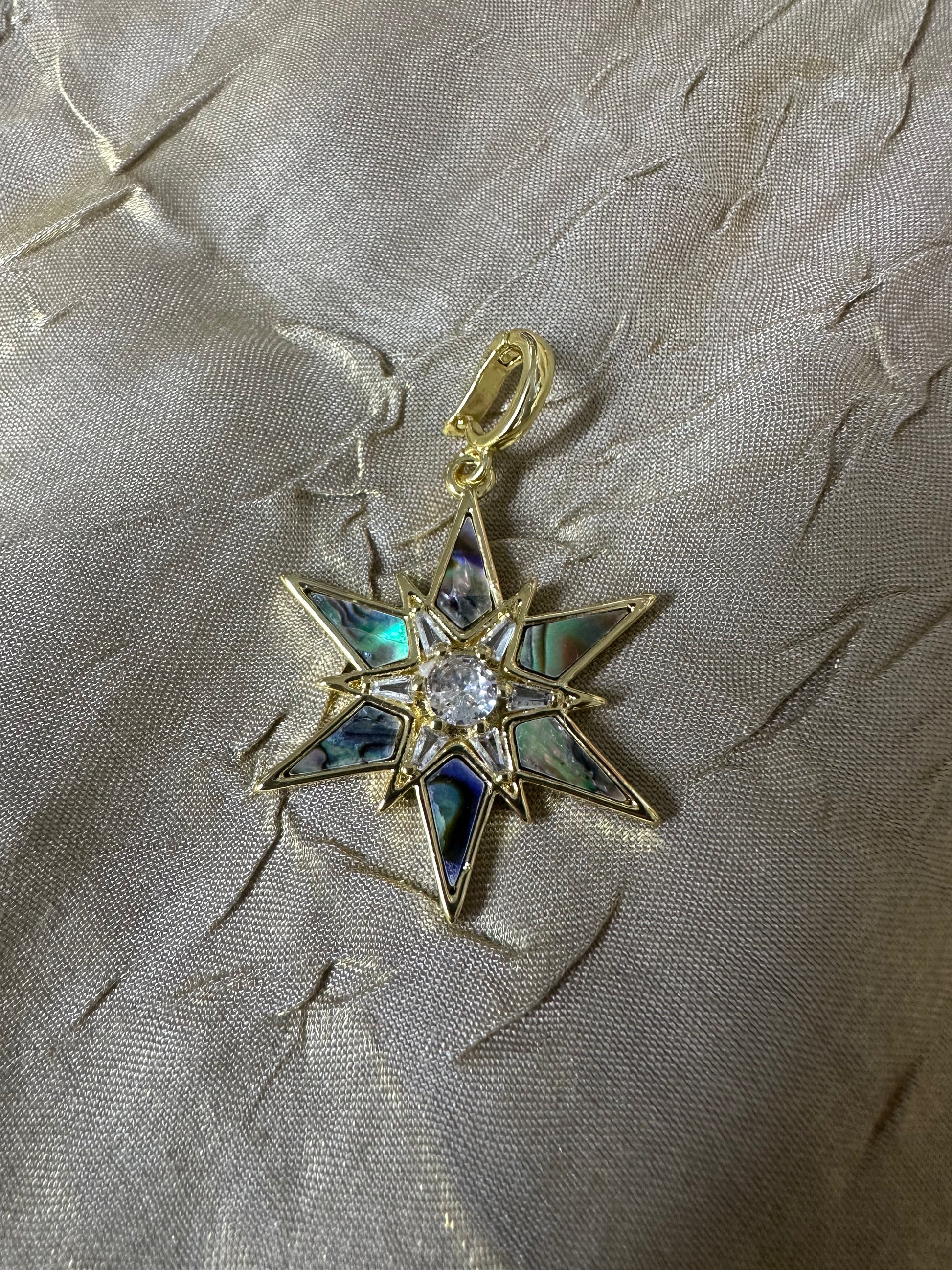 A gold star-shaped pendant with iridescent abalone shell inlays and a clear central stone, displayed on a textured beige fabric background.