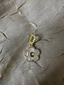 A gold-tone flower-shaped charm with a white mother-of-pearl center and a beaded border, displayed on a textured beige fabric background.