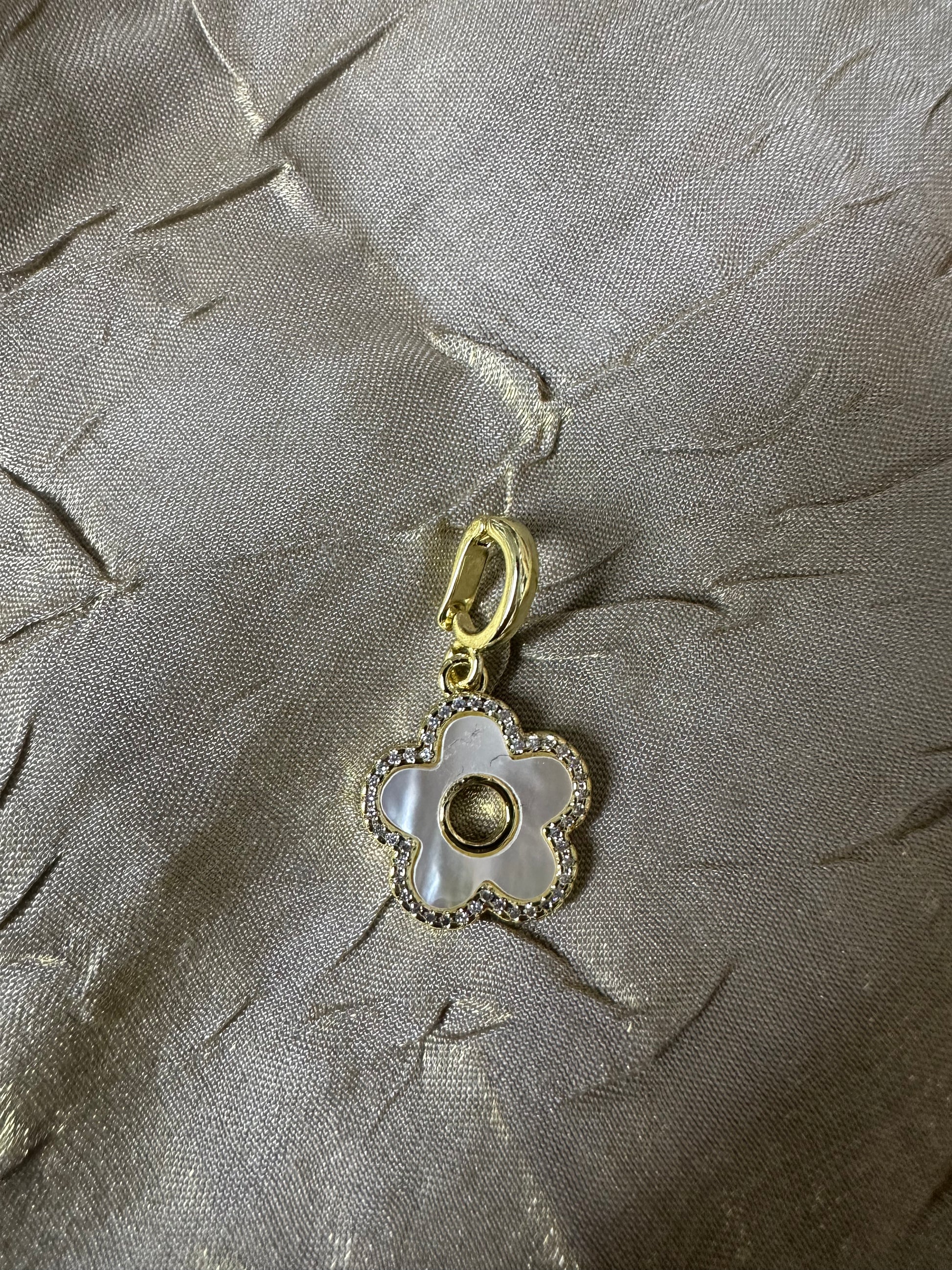 A gold-tone flower-shaped charm with a white mother-of-pearl center and a beaded border, displayed on a textured beige fabric background.