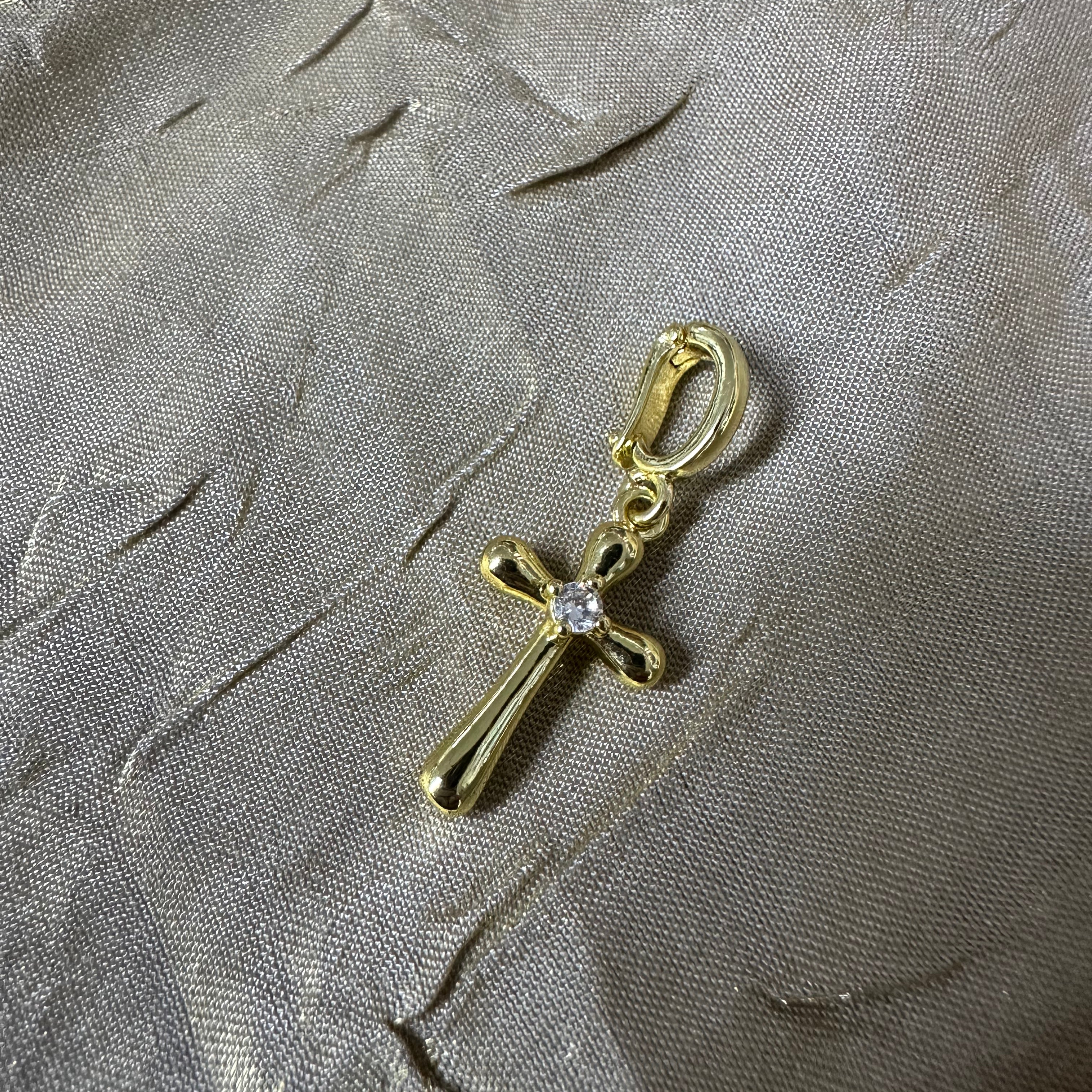 A gold cross charm with a small clear gemstone at the center, attached to a gold lobster clasp, displayed on a textured gray fabric background.