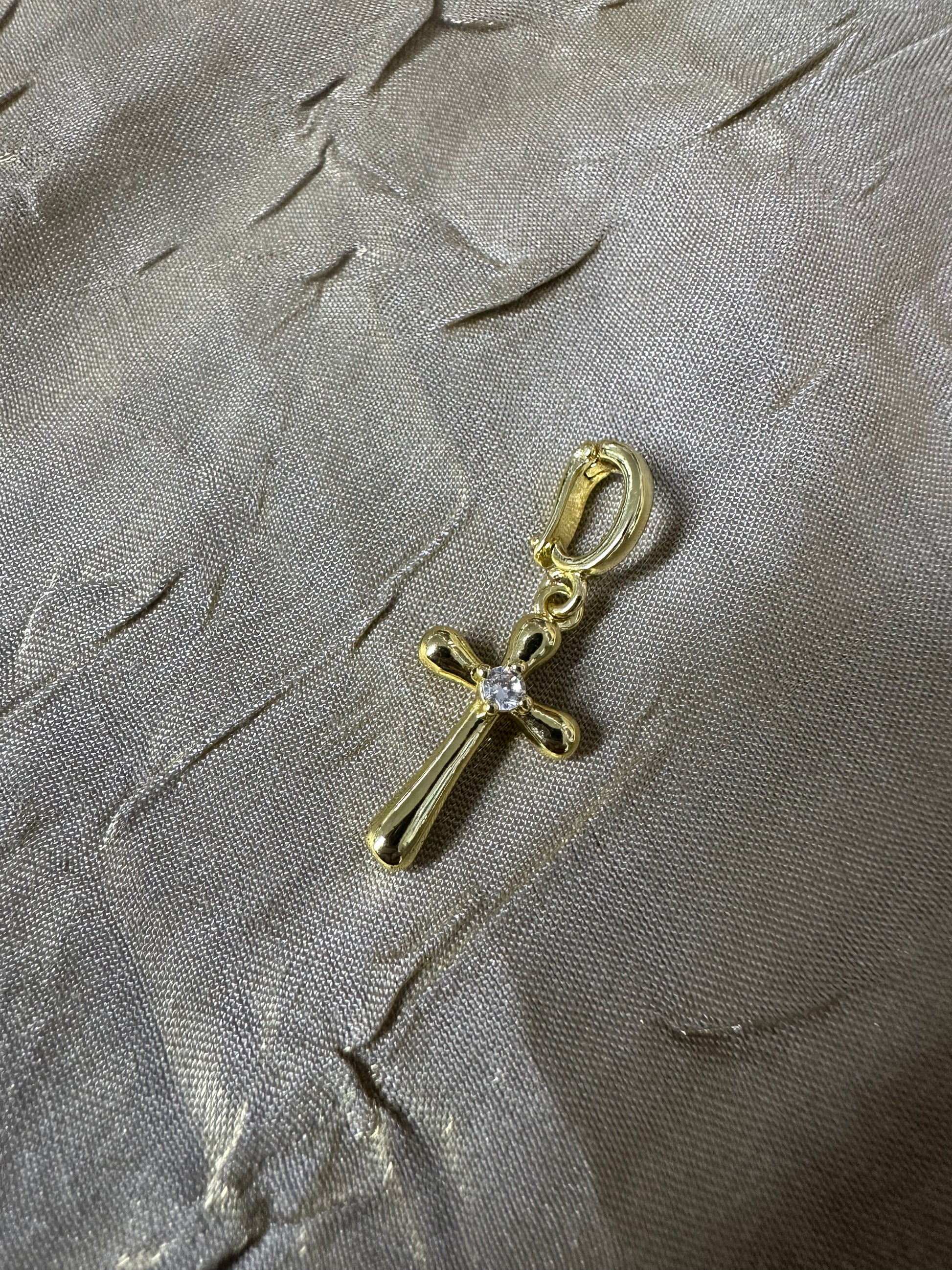 A gold cross charm with a small clear gemstone at the center, attached to a gold lobster clasp, displayed on a textured gray fabric background.