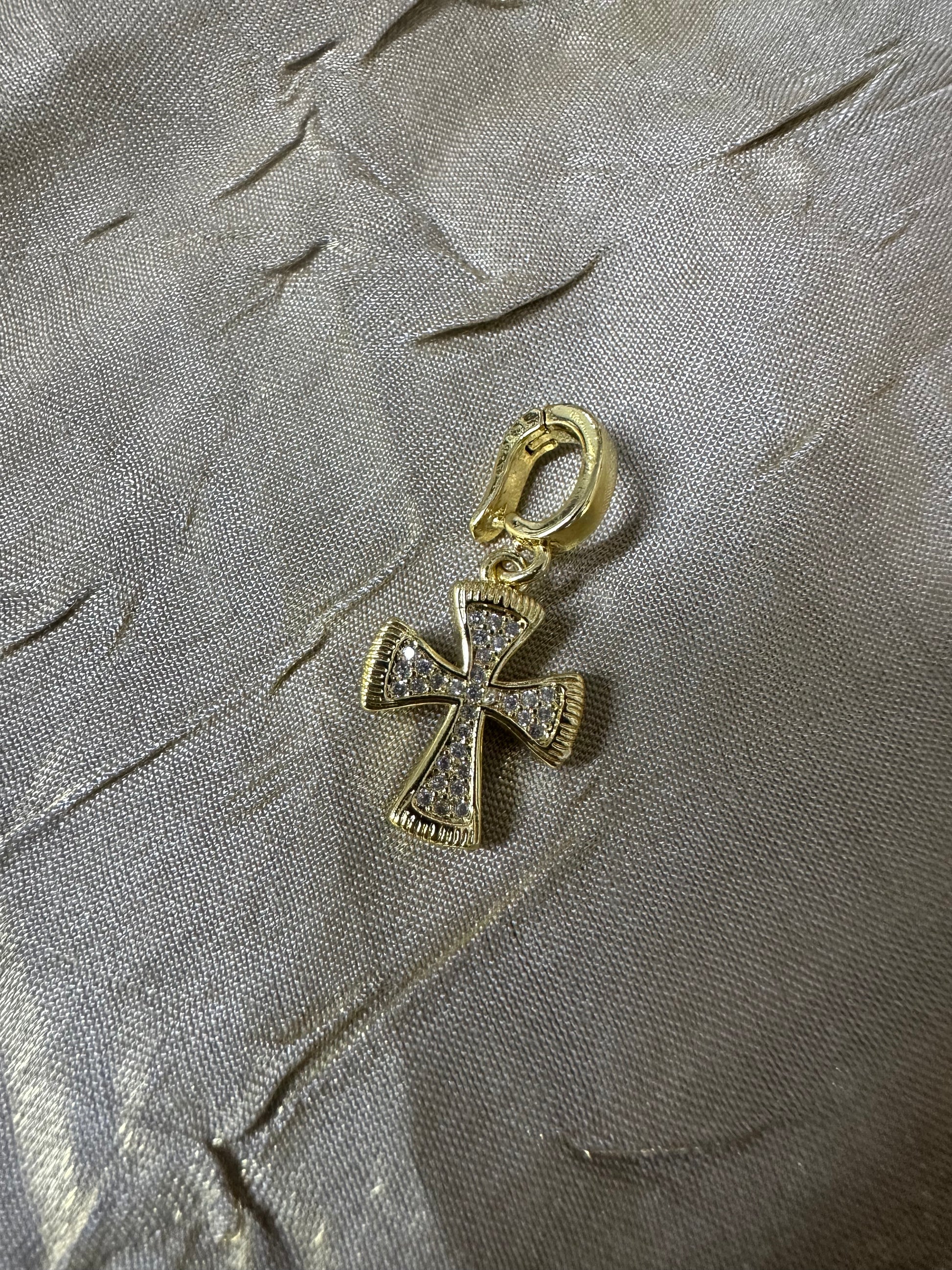 A gold cross pendant charm embellished with clear crystals, resting on a textured silver fabric background.