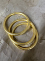 Three textured gold bangles stacked together on a wrinkled gray paper background.