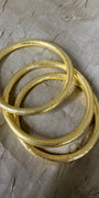 Three textured gold bangles stacked together on a wrinkled gray paper background.