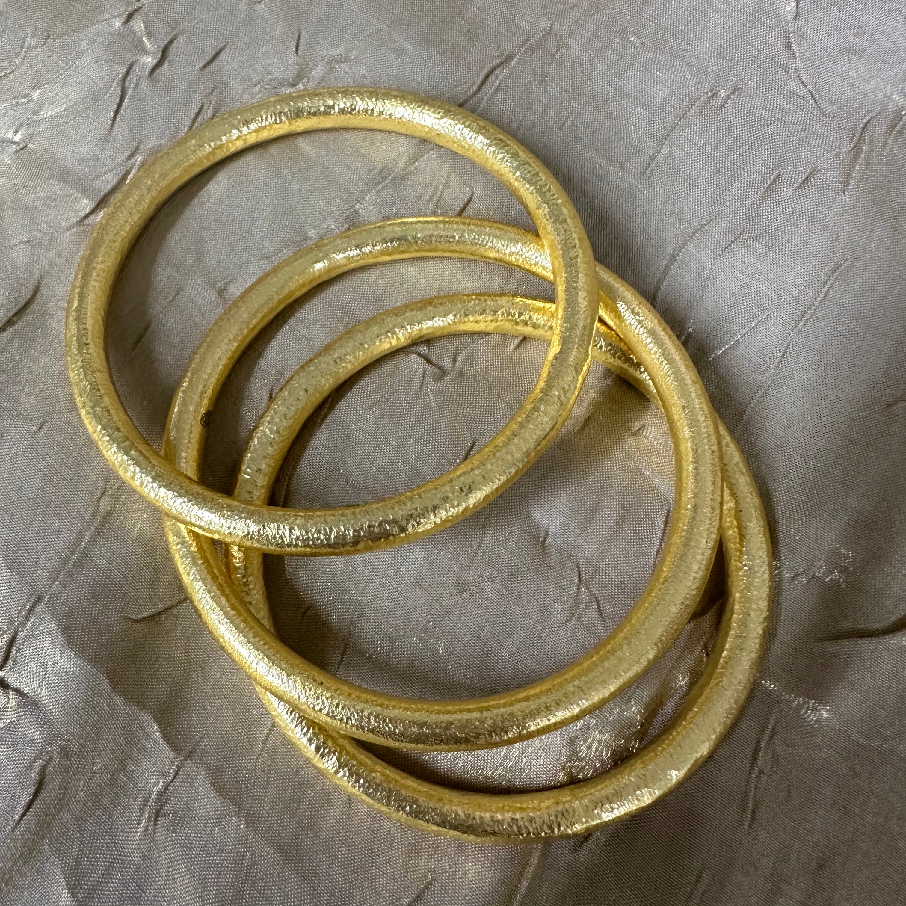 Three textured gold bangles stacked together on a wrinkled gray paper background.