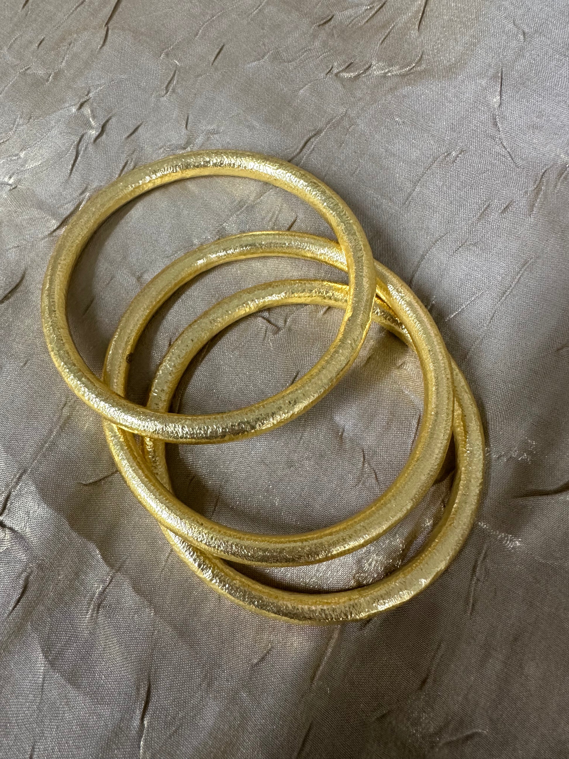 Three textured gold bangles stacked together on a wrinkled gray paper background.