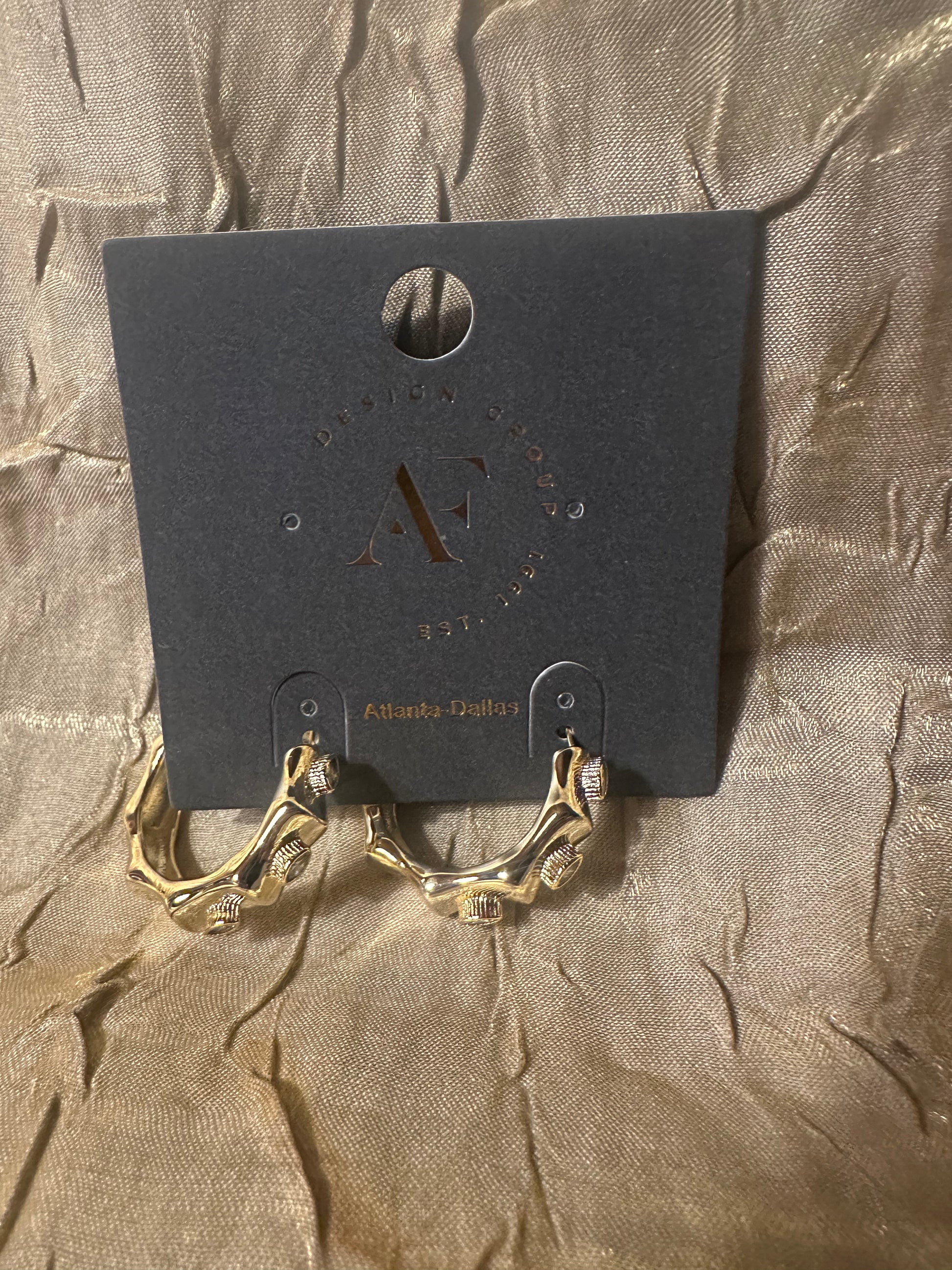 Gold hoop earrings with clear crystal-like accents, displayed on a black branded jewelry card against a textured beige fabric background.