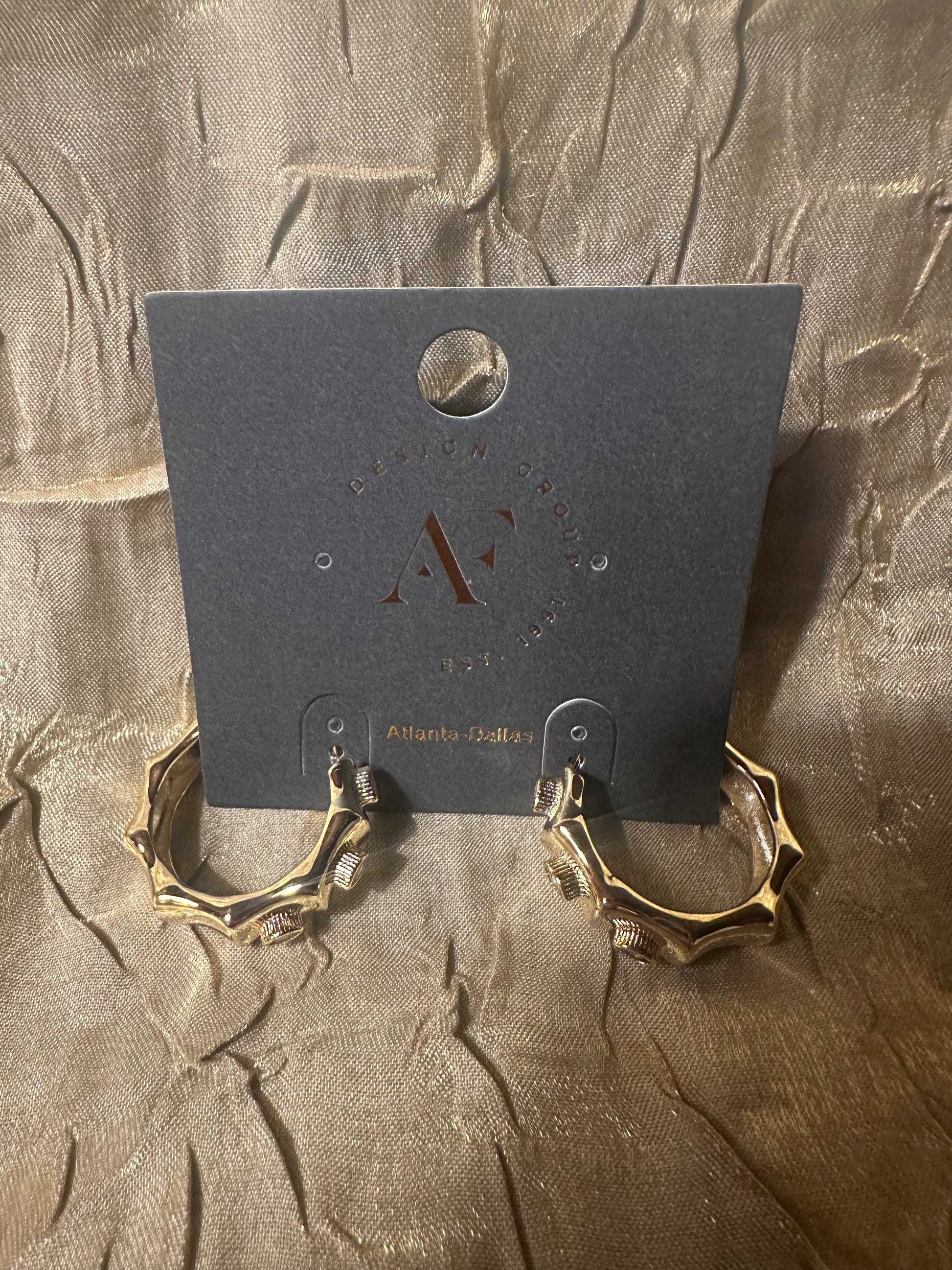 A pair of chunky gold hoop earrings with textured, organic edges, displayed on a dark gray branded card against a crinkled gold fabric background.
