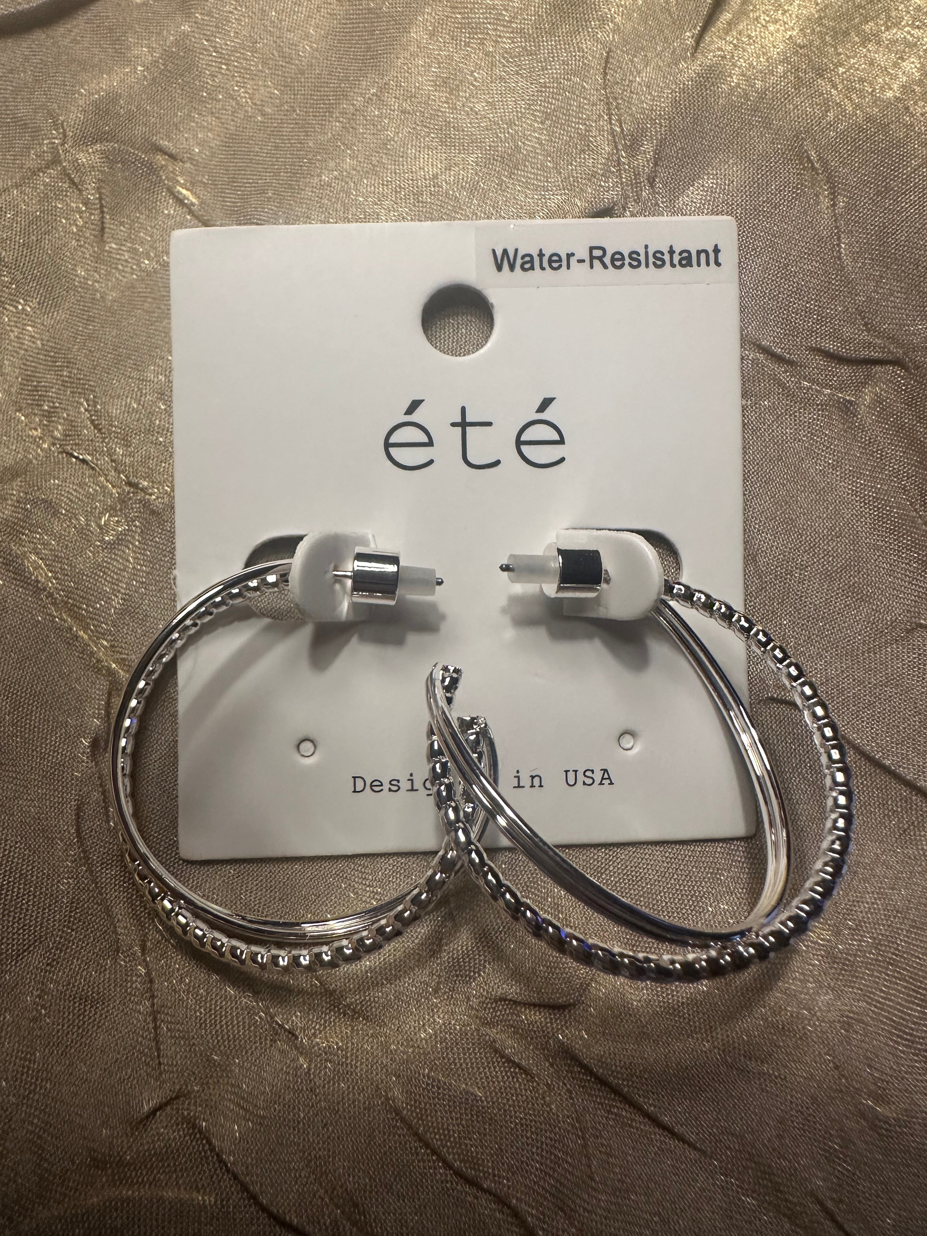 A pair of silver hoop earrings with textured beaded edges and white silicone tips, displayed on a white card labeled 'eté' against a textured gold fabric background.