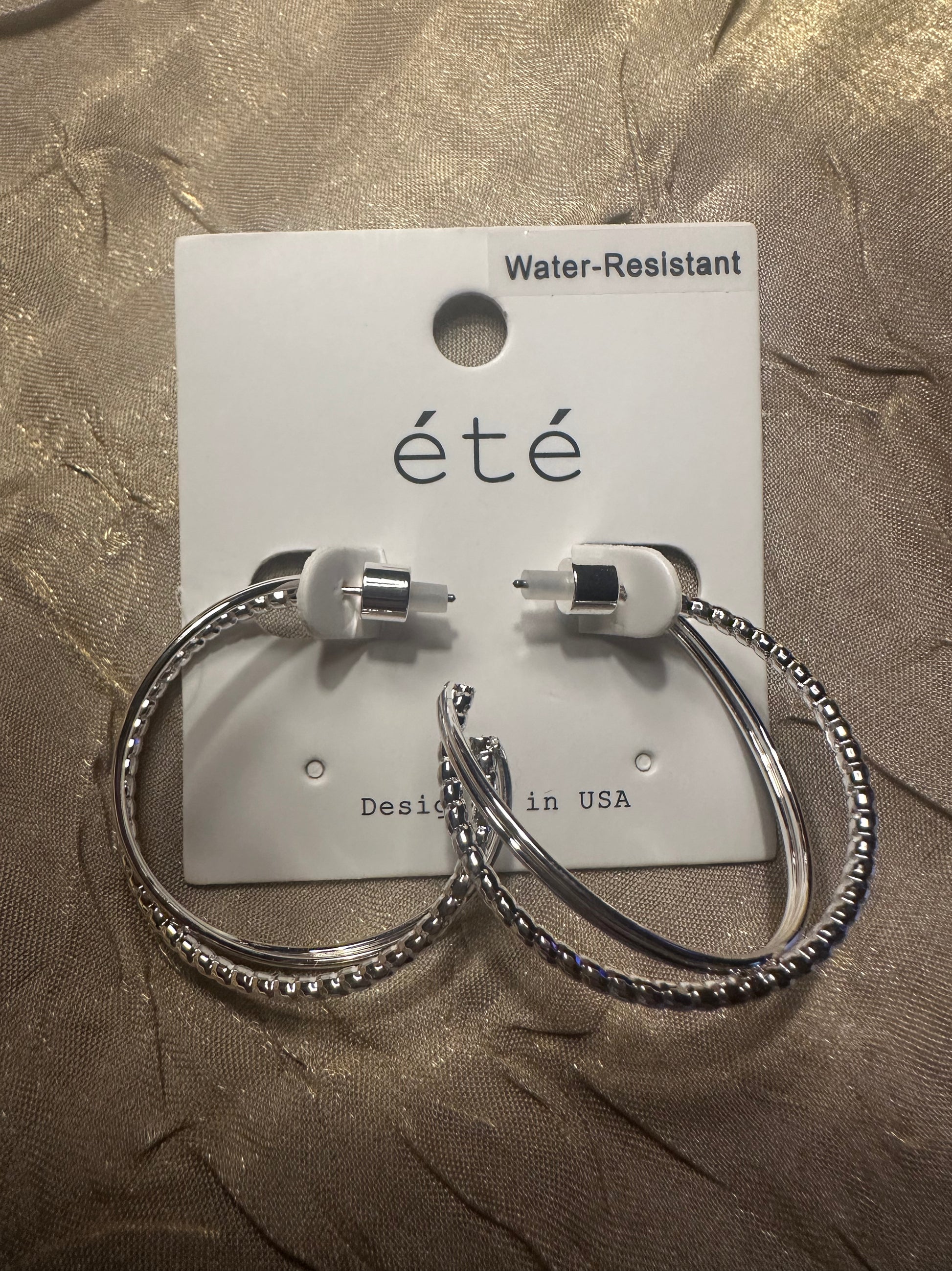 A pair of silver hoop earrings with textured beaded edges and white silicone tips, displayed on a white card labeled 'eté' against a textured gold fabric background.