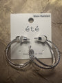 A pair of silver-tone hoop earrings with textured beaded edges, displayed on a white card labeled “été” and “Water-Resistant,” placed on a textured metallic gold surface.