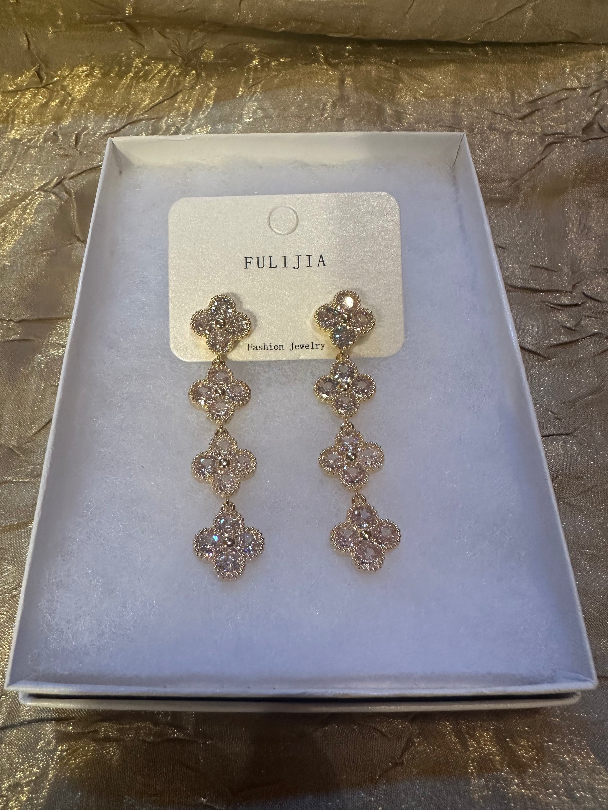 A pair of gold-tone drop earrings with clear rhinestones in a four-flower clover design, presented on a white card inside a white jewelry box. The brand name 'FULIJIA' is visible on the card.