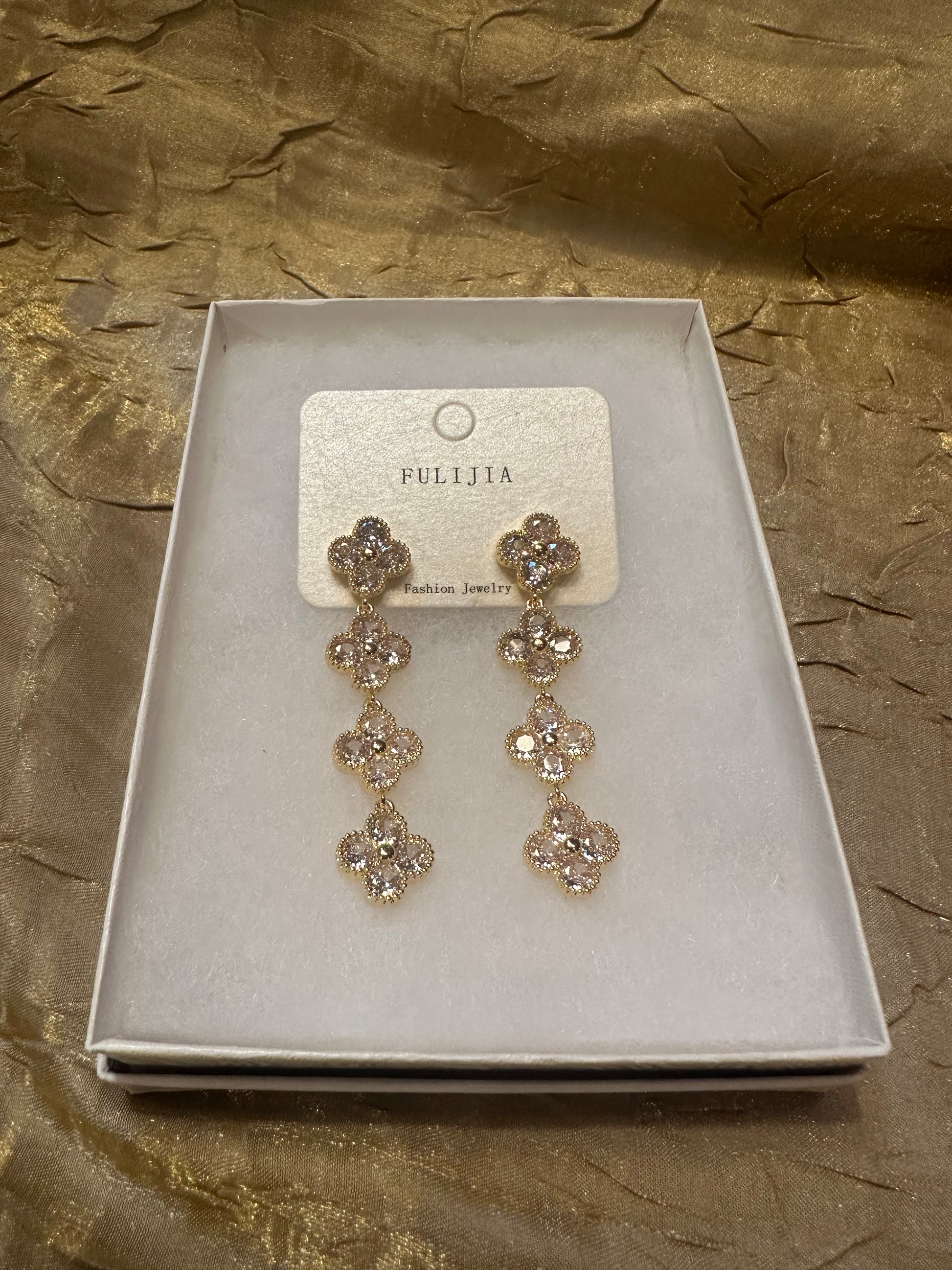 A pair of gold-toned dangle earrings with clear rhinestones, presented on a white jewelry card inside a white box, set against a textured gold fabric background. The brand name 'FULIJA' is visible on the card.