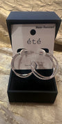 A pair of large silver hoop earrings with clear rhinestone-like stones, displayed in an open black jewelry box with a white card labeled “été” and “Water-Resistant.”.