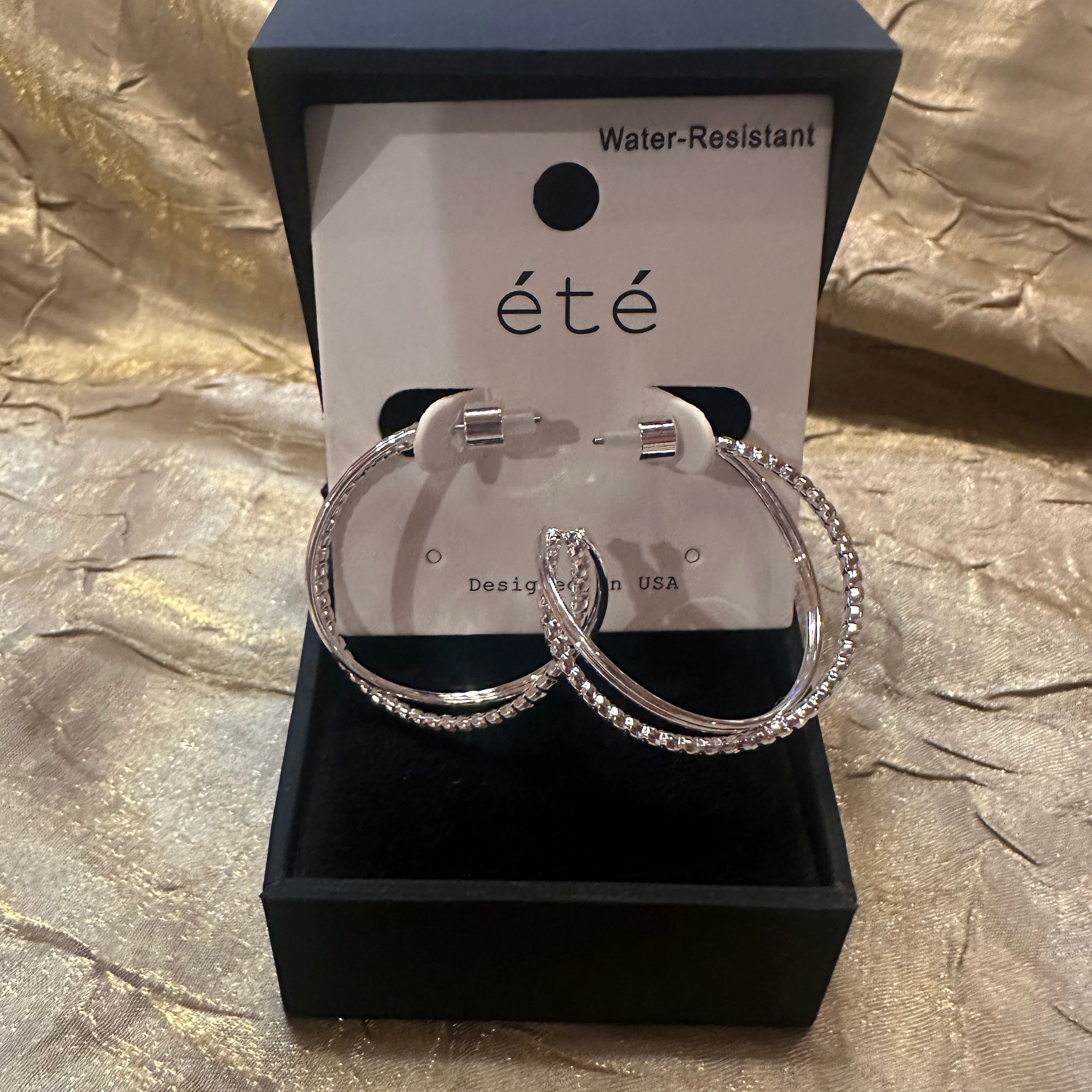 A pair of large silver hoop earrings with clear rhinestone-like stones, displayed in an open black jewelry box with a white card labeled “été” and “Water-Resistant.”.