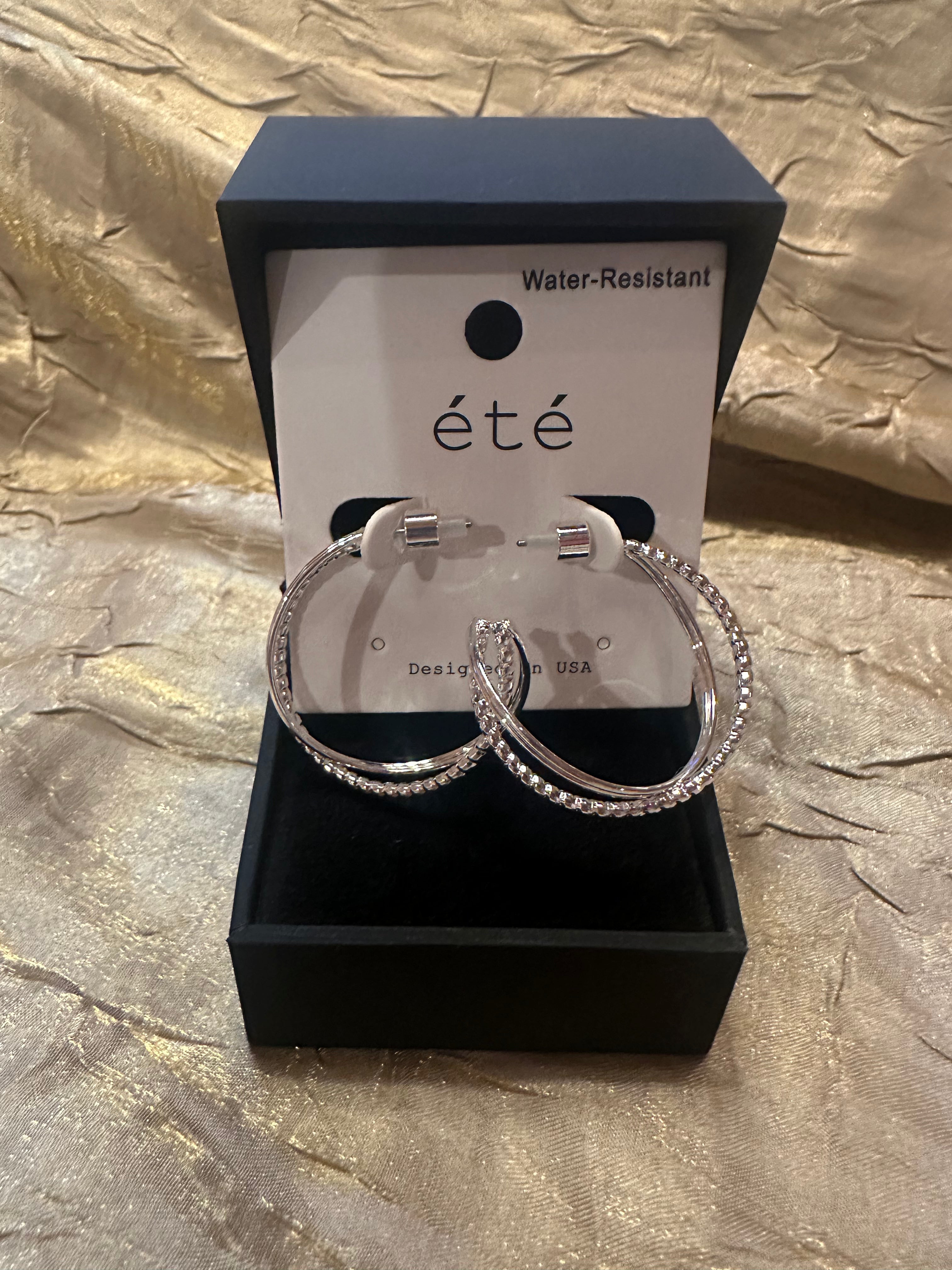 A pair of large silver hoop earrings with clear rhinestone-like stones, displayed in an open black jewelry box with a white card labeled “été” and “Water-Resistant.”.