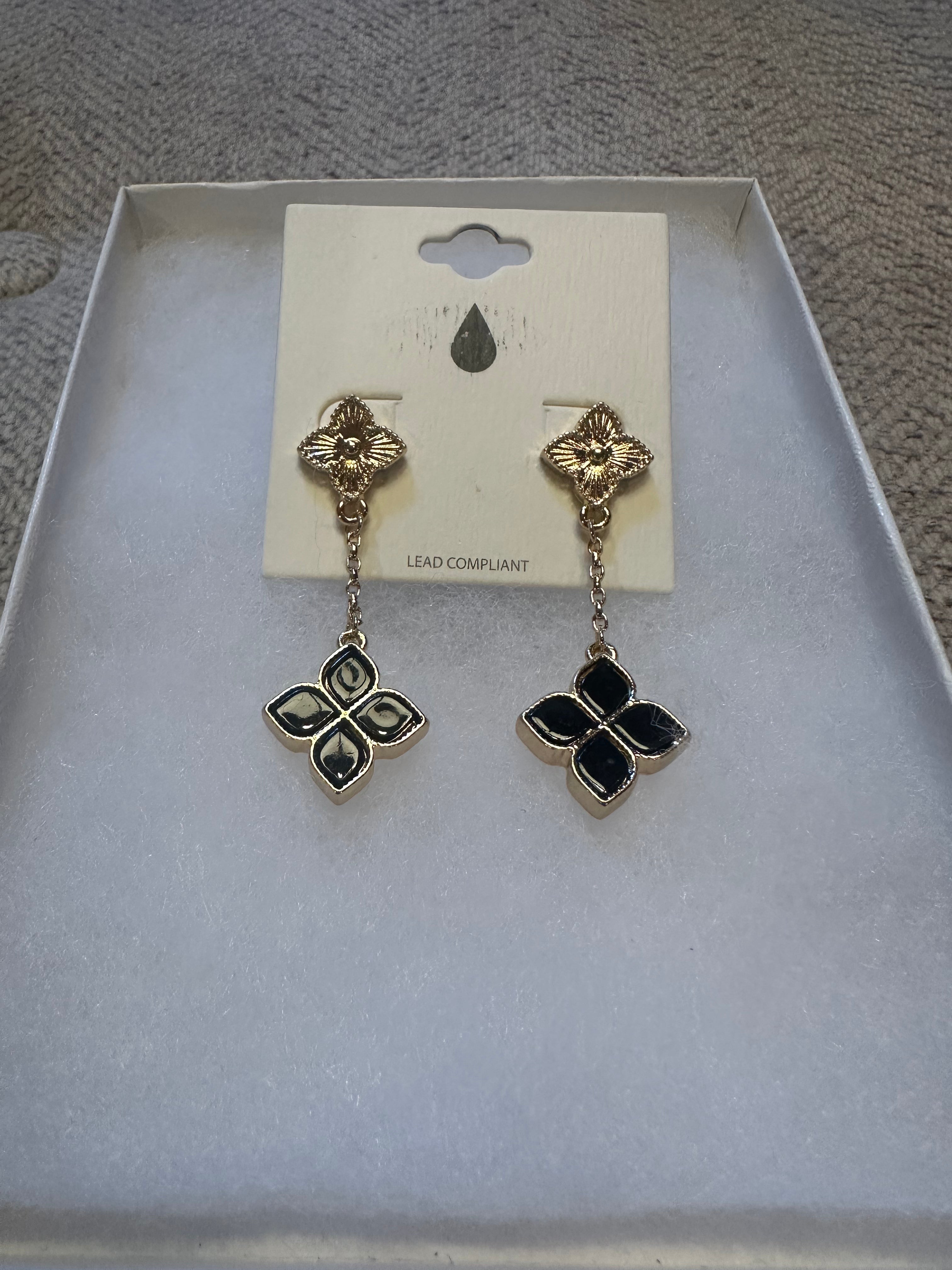A pair of gold-tone drop earrings with black enamel clover-shaped charms, displayed on a white card inside a white jewelry box.