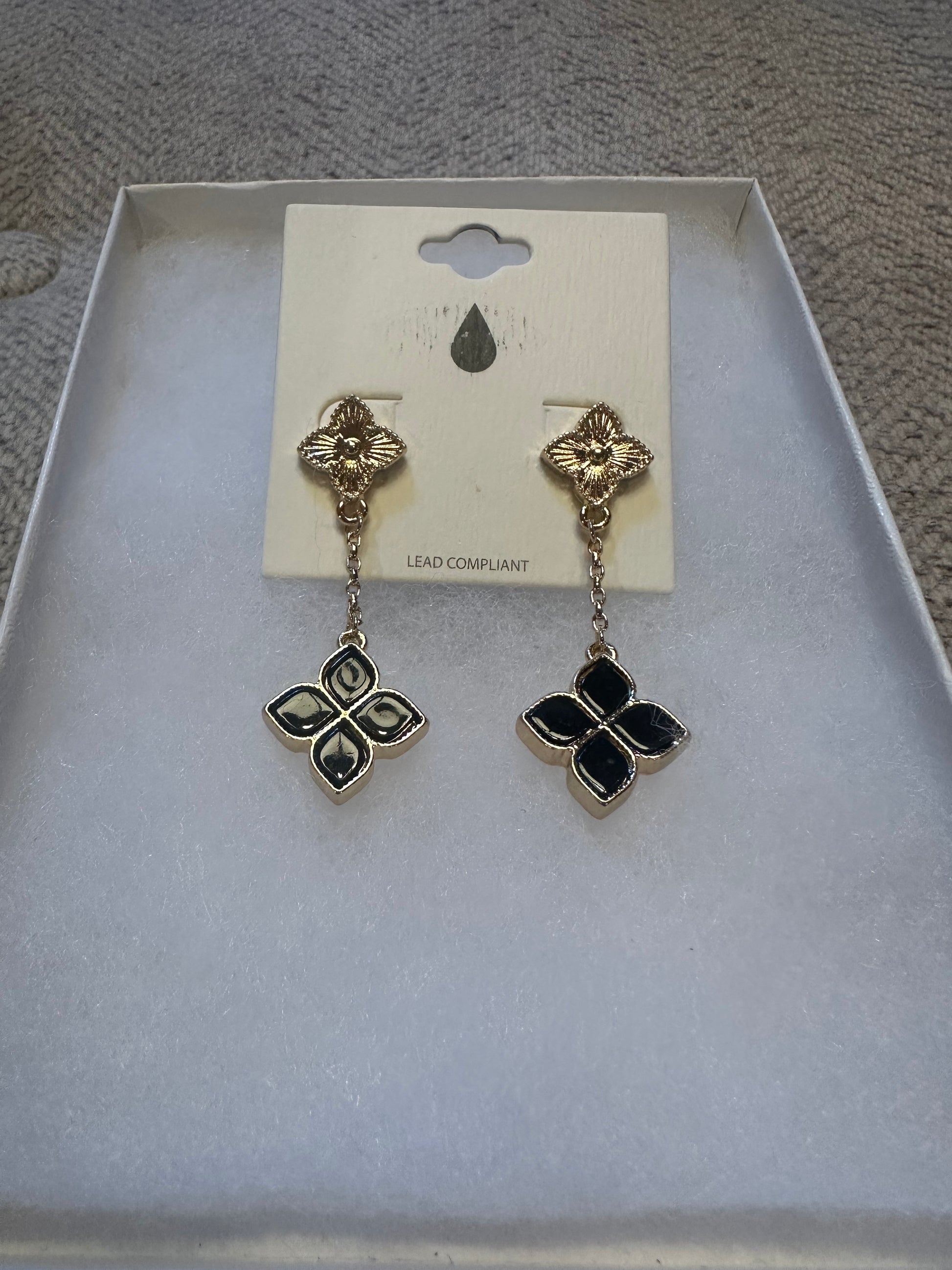 A pair of gold-tone drop earrings with black enamel clover-shaped charms, displayed on a white card inside a white jewelry box.