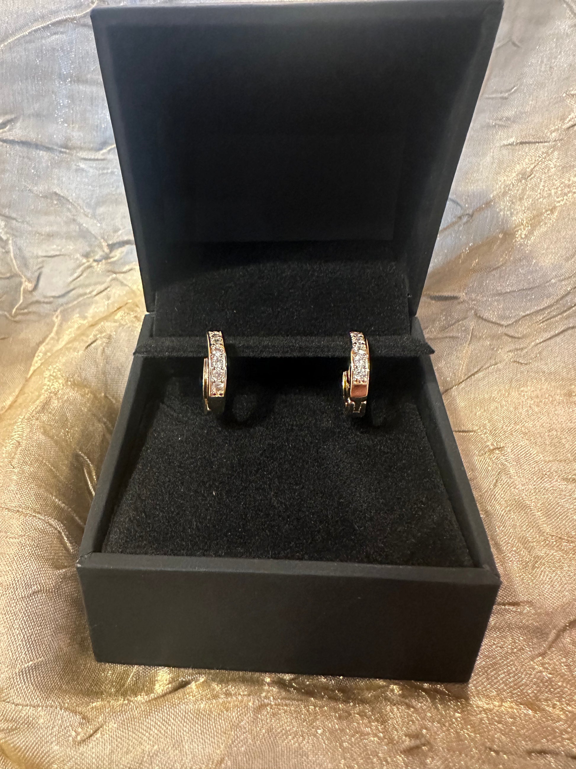 A pair of gold hoop earrings with pavé-set clear stones, displayed inside an open black jewelry box on a textured gold fabric background.