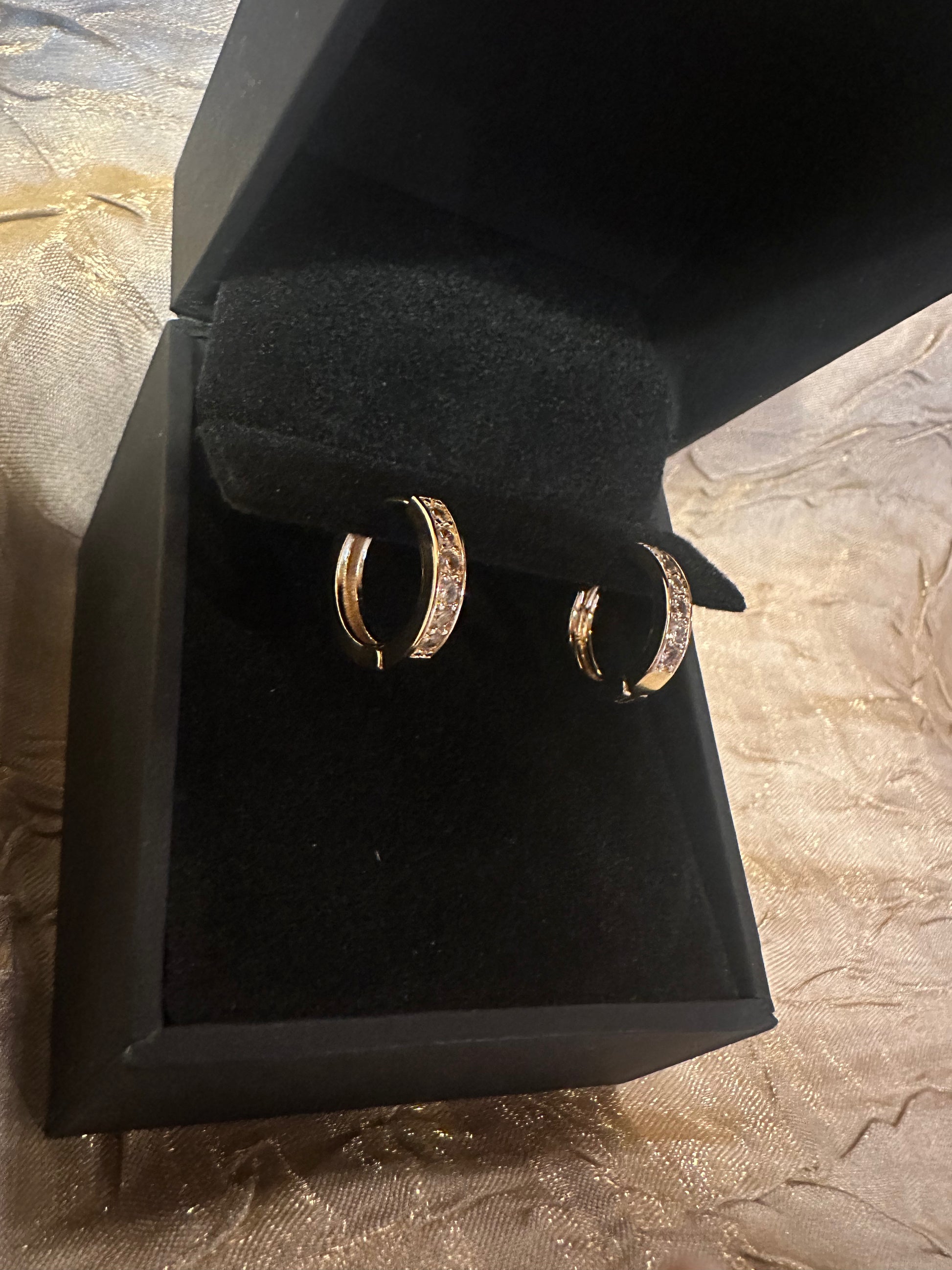 A pair of gold hoop earrings with small clear stones, displayed inside an open black jewelry box on a textured beige fabric background.
