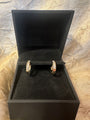 A pair of gold hoop earrings with clear pavé stones, displayed in an open black jewelry box on a textured gold fabric background.