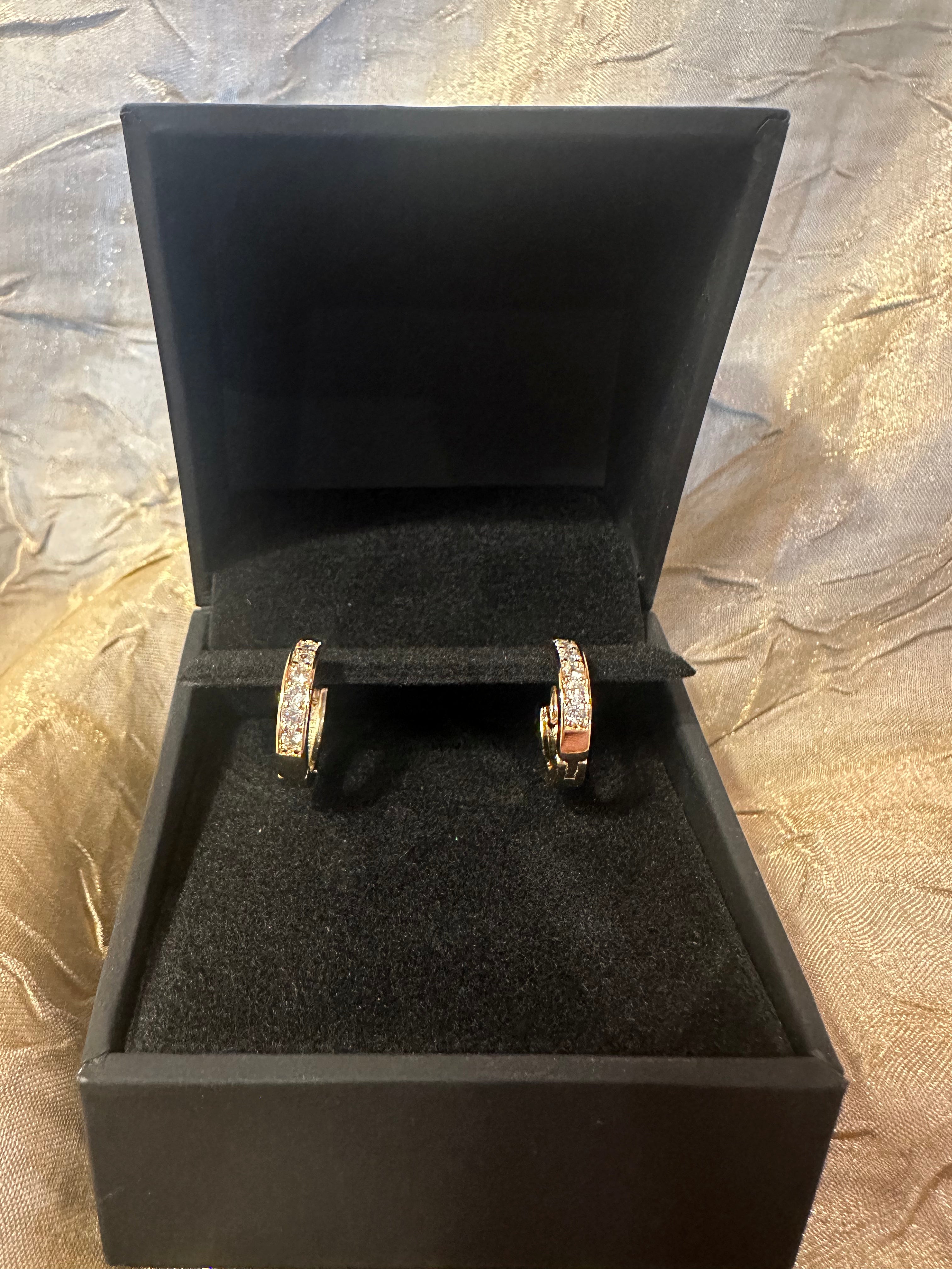 A pair of gold hoop earrings with clear pavé stones, displayed in an open black jewelry box on a textured gold fabric background.