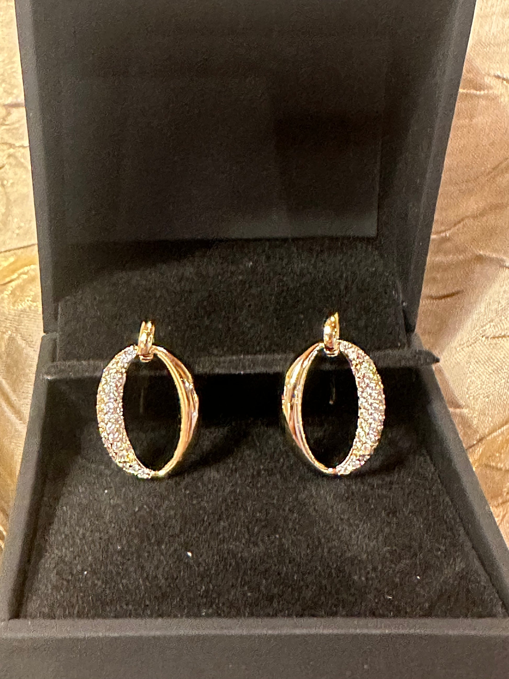 A pair of gold hoop earrings with pavé-set clear stones on the front curves, displayed inside an open black jewelry box.