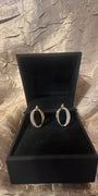 A pair of gold hoop earrings with pavé-set clear stones, displayed inside an open black jewelry box on a textured metallic gold fabric background.