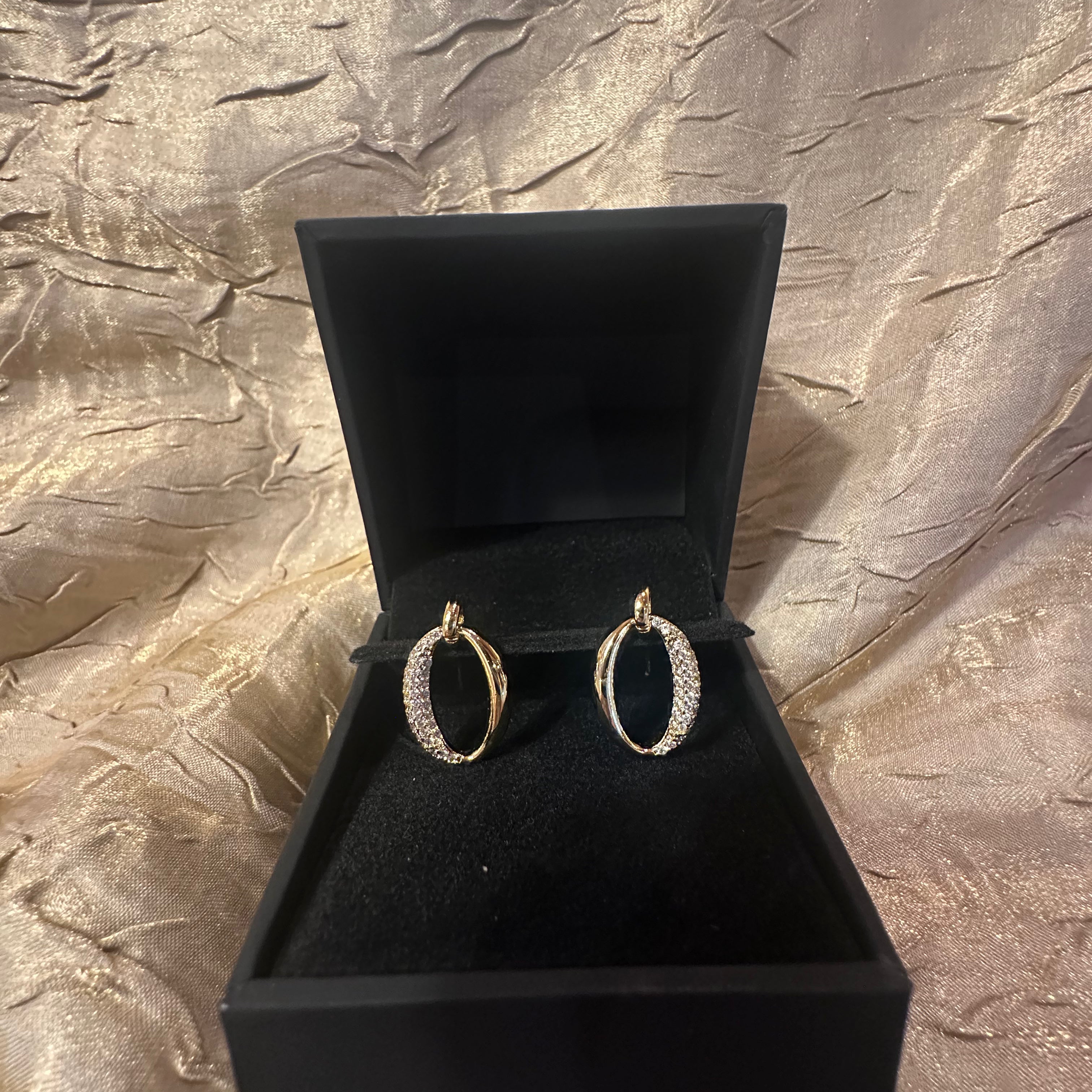 A pair of gold hoop earrings with pavé-set clear stones, displayed inside an open black jewelry box on a textured metallic gold fabric background.