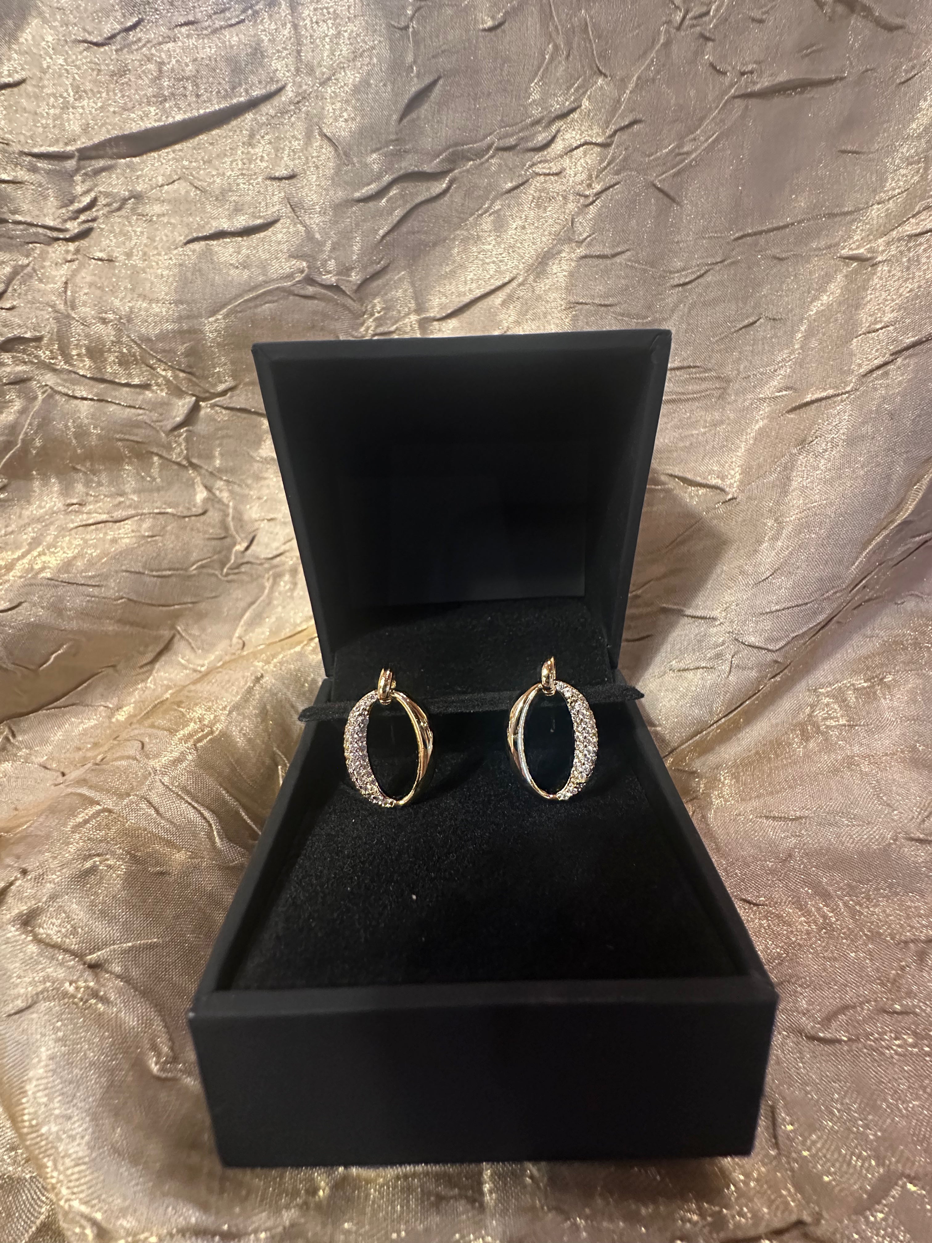 A pair of gold hoop earrings with pavé-set clear stones, displayed inside an open black jewelry box on a textured metallic gold fabric background.