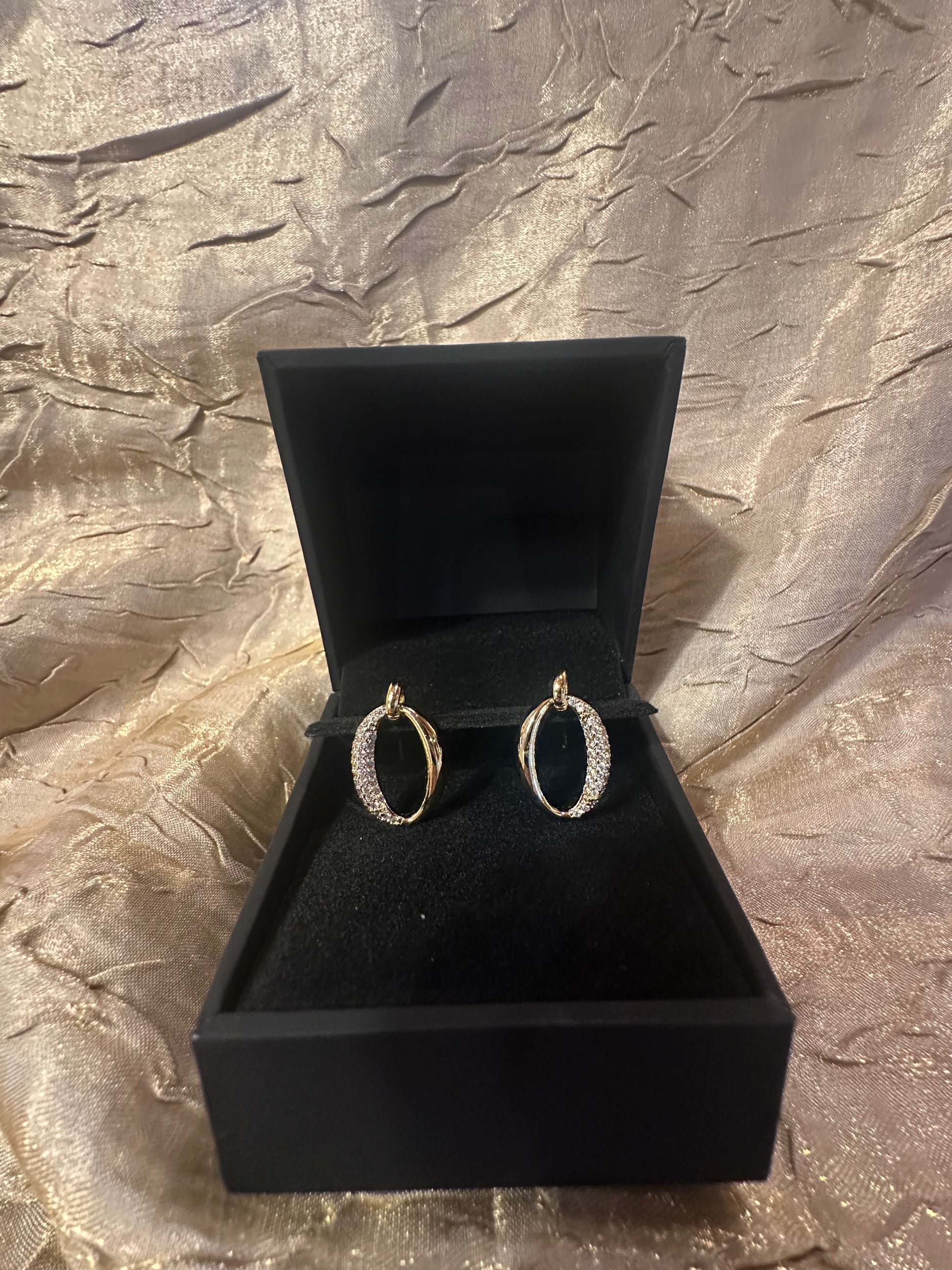 A pair of gold hoop earrings with pavé-set clear stones, displayed inside an open black jewelry box on a textured metallic gold fabric background.