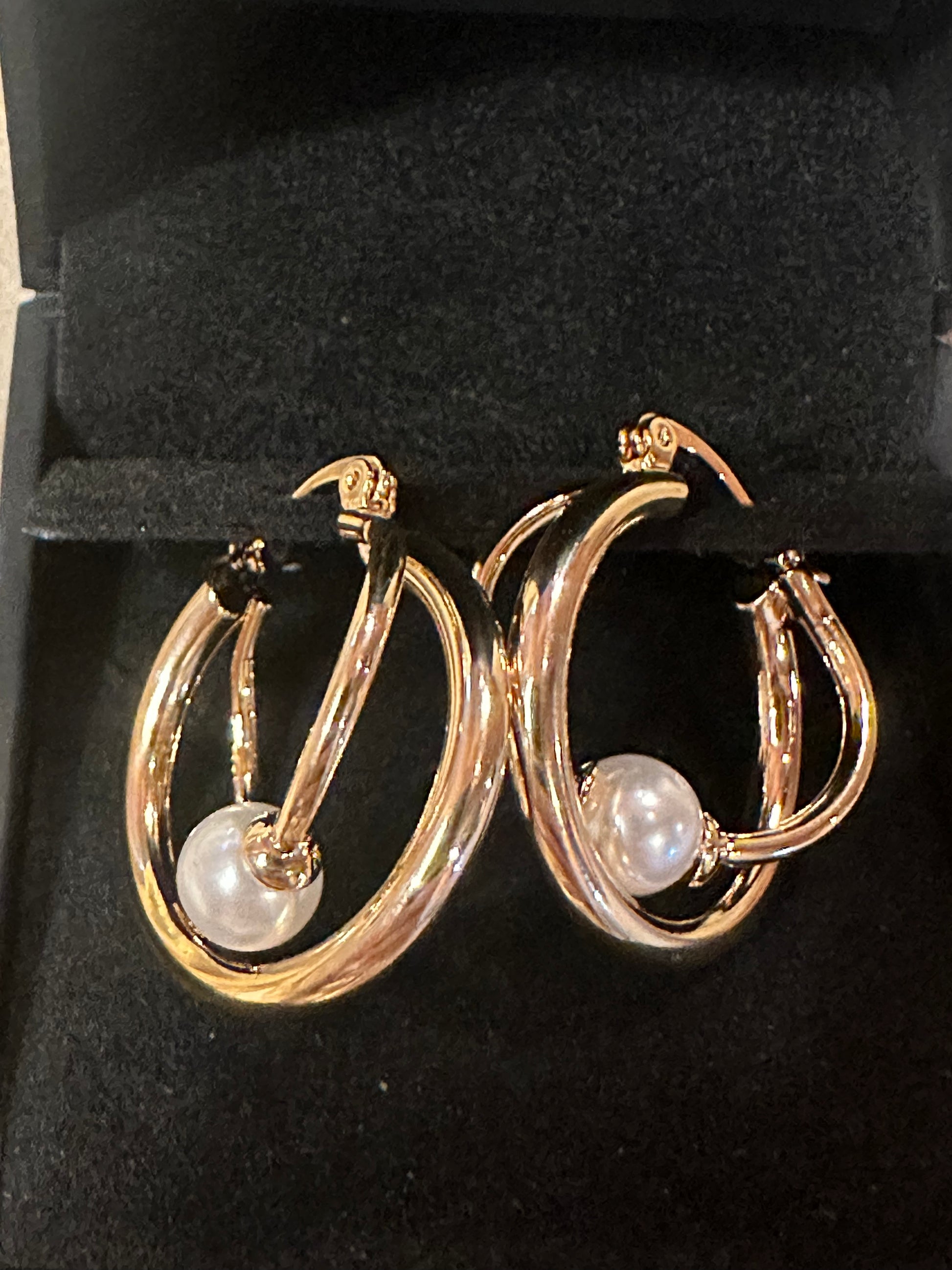 A pair of gold hoop earrings with a white pearl dangling from each hoop, displayed inside a black jewelry box.