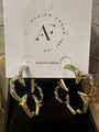 A pair of gold-toned abstract hoop earrings with clear rhinestone accents, displayed on a black jewelry box insert in front of a white hanging card printed with the brand name 'Design Group Atlanta-Dallas'.