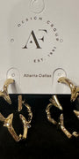 A pair of gold-toned abstract hoop earrings with clear rhinestone accents, displayed on a black jewelry box insert in front of a white hanging card printed with the brand name 'Design Group Atlanta-Dallas'.