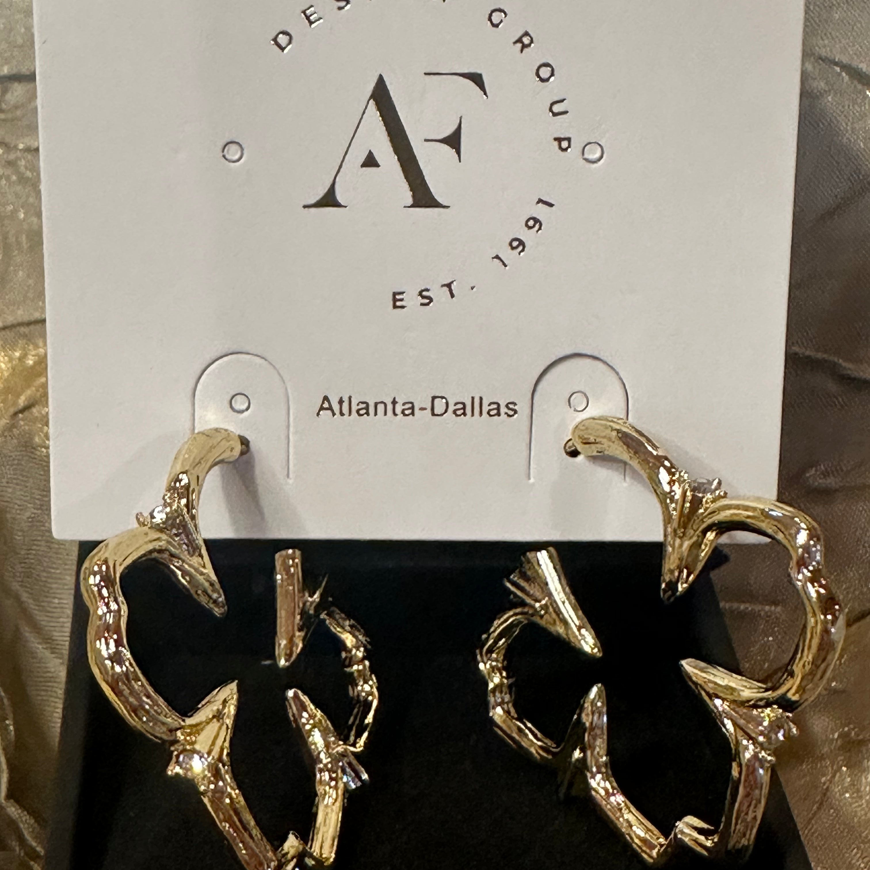 A pair of gold-toned abstract hoop earrings with clear rhinestone accents, displayed on a black jewelry box insert in front of a white hanging card printed with the brand name 'Design Group Atlanta-Dallas'.