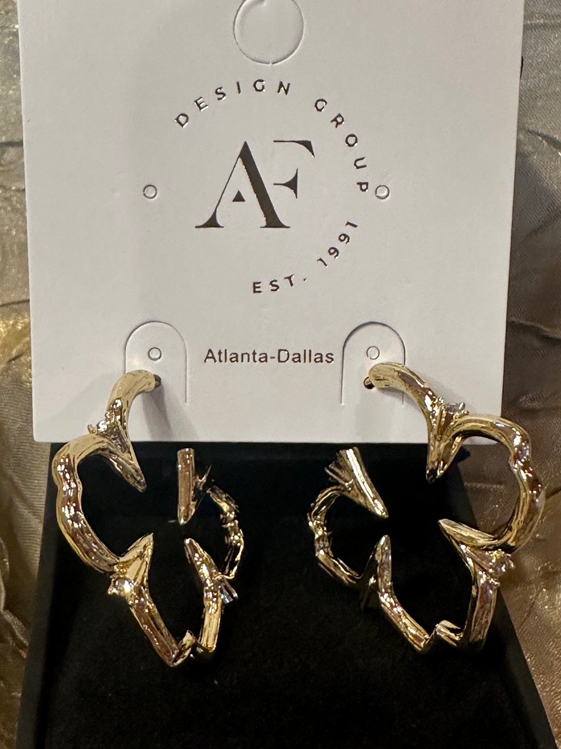A pair of gold-toned abstract hoop earrings with clear rhinestone accents, displayed on a black jewelry box insert in front of a white hanging card printed with the brand name 'Design Group Atlanta-Dallas'.