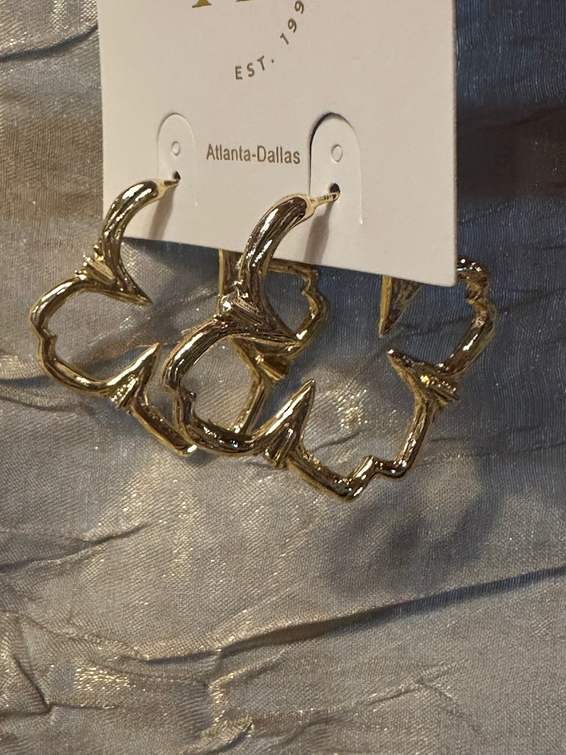 Gold hoop earrings with a textured, organic branch-like design, displayed on a white retail card labeled 'Atlanta-Dallas' against a shimmering silver fabric background.