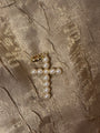 A gold cross pendant embellished with white faux pearls, resting on a textured, shimmering champagne-colored fabric background.